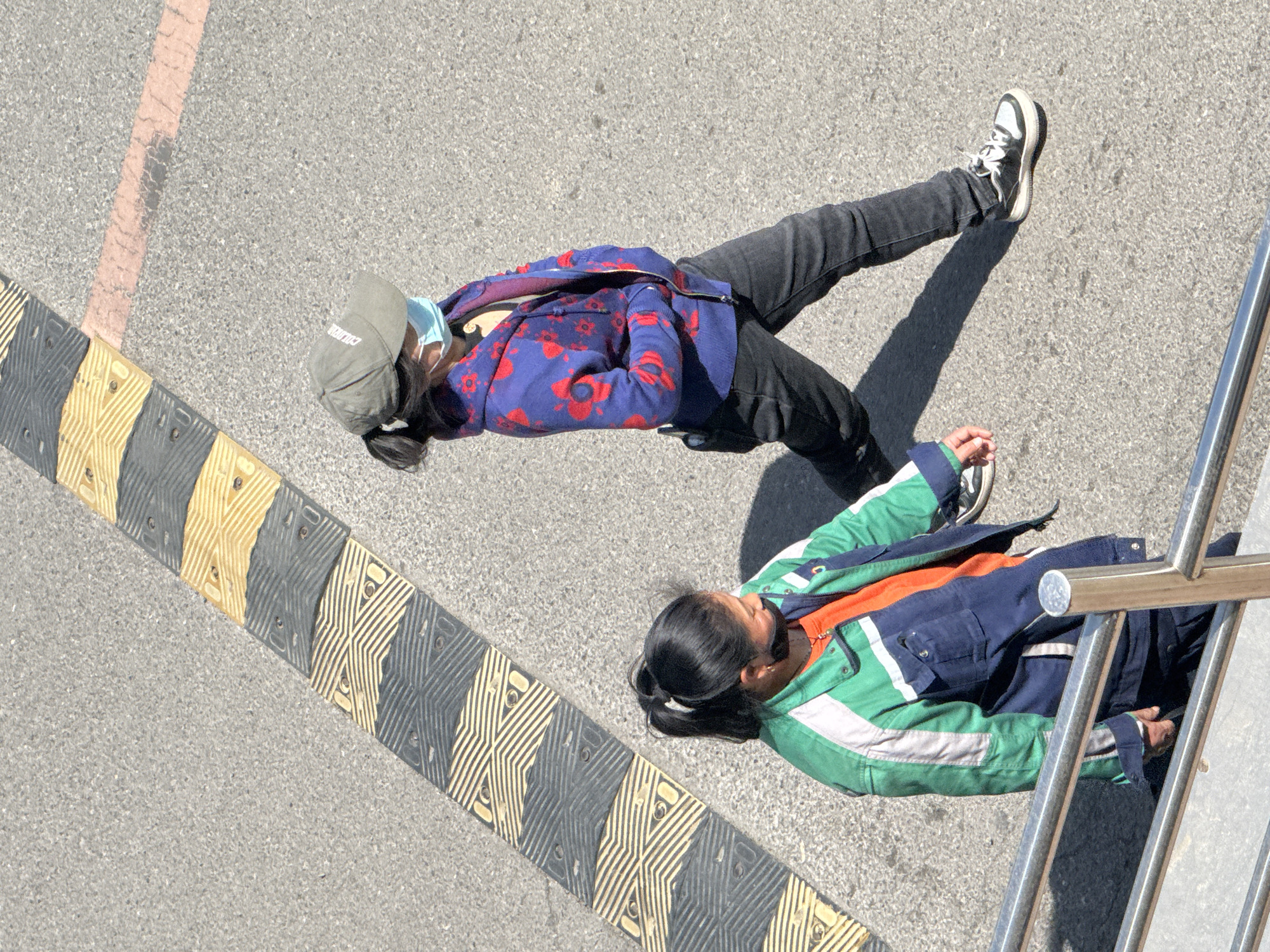 Two people lie on an asphalt surface near a speed bump and metal railing. The first person, wearing black pants, a dark blue jacket with red floral patterns, a gray hat, and a light blue face mask, is positioned next to a speed bump marked with yellow and black diagonal stripes. The second person, wearing a dark blue and green jacket with an orange shirt underneath, is lying next to the metal railing. The location is Zhongtun, China.