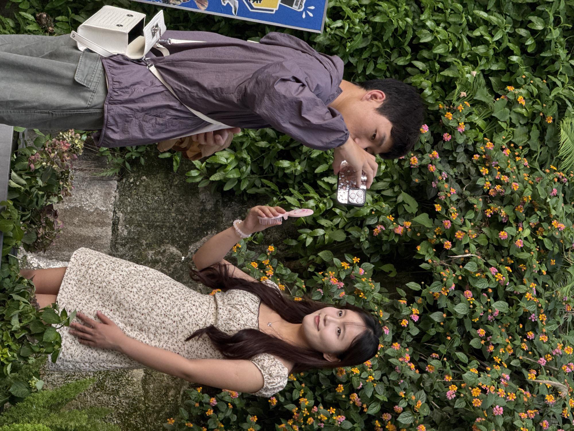 In Yuzhong District, China, a young man wearing a maroon windbreaker and grey pants is taking a picture of a young woman in a floral dress. The woman is holding a pink comb and posing. They are surrounded by greenery and flowers, with a stone pathway visible nearby. The man has a white purse slung over his shoulder.