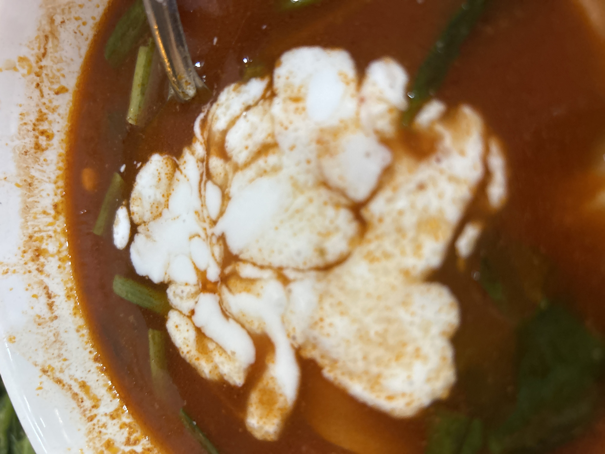 A close-up shot of a bowl of Vietnamese soup, potentially Bún cá Nha Trang, with a white swirl of topping (possibly coconut milk or cream) in the center. Green vegetable pieces are visible within the reddish-brown broth. A metal spoon rests near the left edge of the bowl, which appears to be white ceramic. The scene suggests a food setting, likely in the city of Nha Trang, Vietnam, based on the dish. The soup appears to be in a bowl, and it has creamy white topping.
