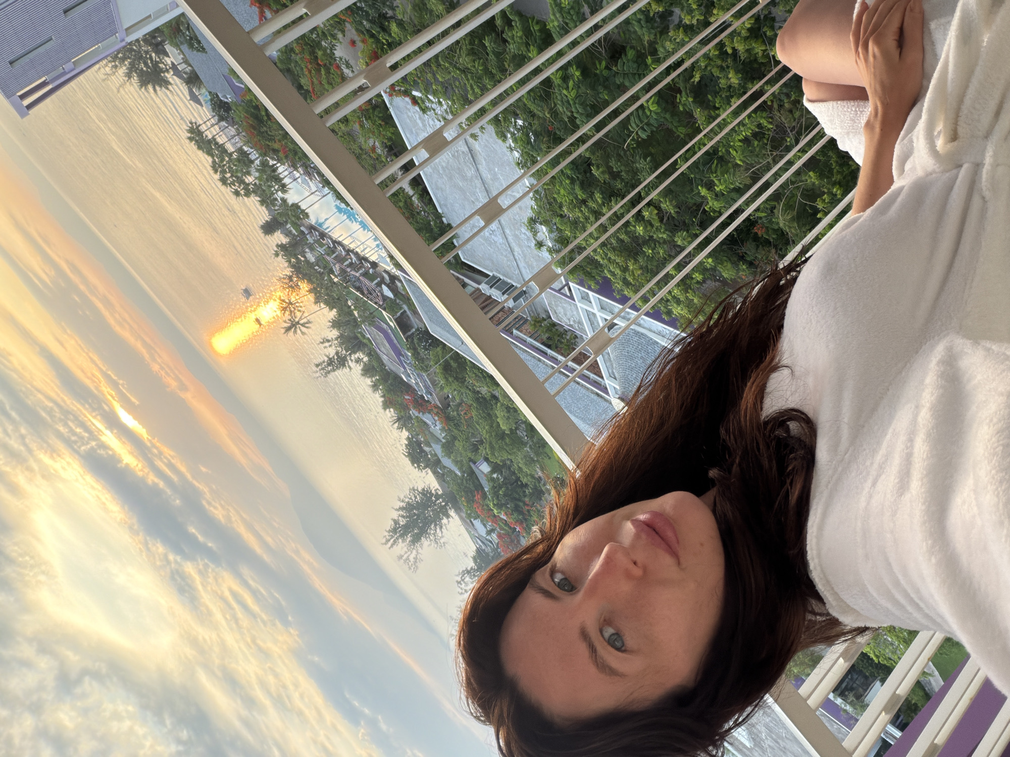 The image shows a woman in a white robe sitting on a balcony, facing the camera. She has long, dark hair and blue eyes. The background shows a sunset over the ocean, with palm trees and buildings visible on the shore. The location is identified as Dong Ninh Hoa Ward, Vietnam. The scene appears to be a resort or hotel setting, with a swimming pool and lounge chairs visible in the distance. The overall mood is relaxed and peaceful.