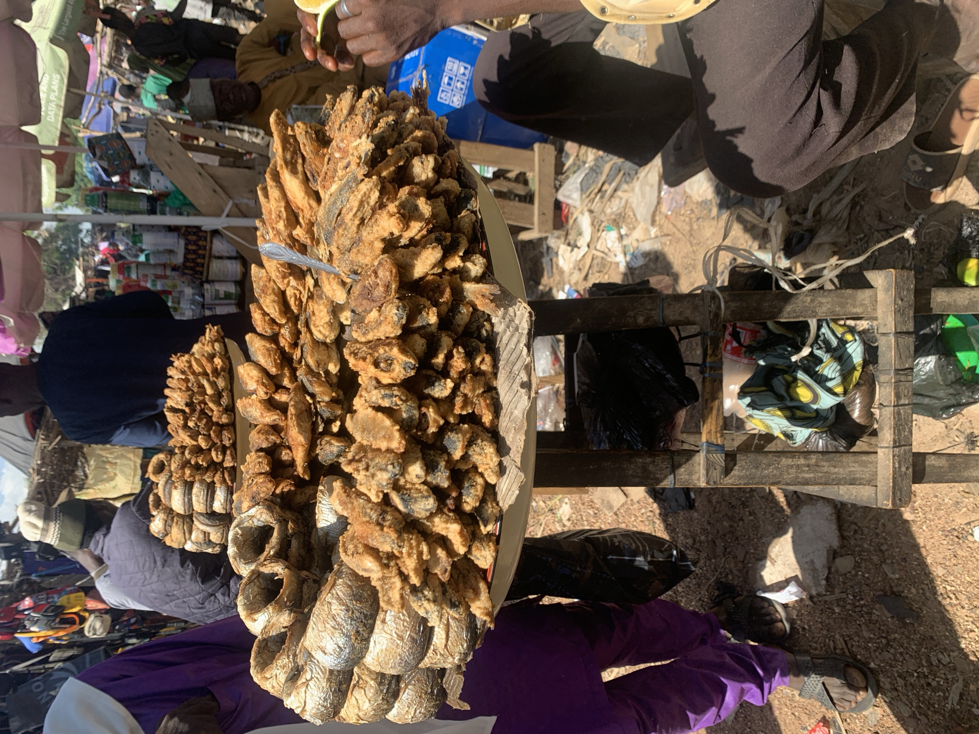 This is a horizontal, eye-level photograph taken outdoors in what appears to be a market setting. The foreground is dominated by a large platter filled with fried fish. The fish are small, whole, and covered in a golden-brown batter, some piled high and others arranged in a circular fashion on the platter. A silver serving spoon is stuck into the fish.
Behind the platter of fish, a wooden stand holds more items, including what looks like more fried fish, bundles of dried fish, and bags of merchandise. The ground around the stand is dusty and littered with debris.
Several people are visible in the background, dressed in various types of clothing. One person's hands are visible in the upper part of the frame, holding a slice of lime and wearing a silver ring. Another person, wearing a dark blue top and a head covering, is partially visible to the left. Further back, several other figures are present, some in more colorful attire. The scene suggests a bustling market atmosphere, likely during daylight hours, given the strong shadows.
The overall impression is a vibrant, active marketplace in Nigeria, focusing on the sale of food, specifically fried fish, and other goods. There is no visible text in the image.