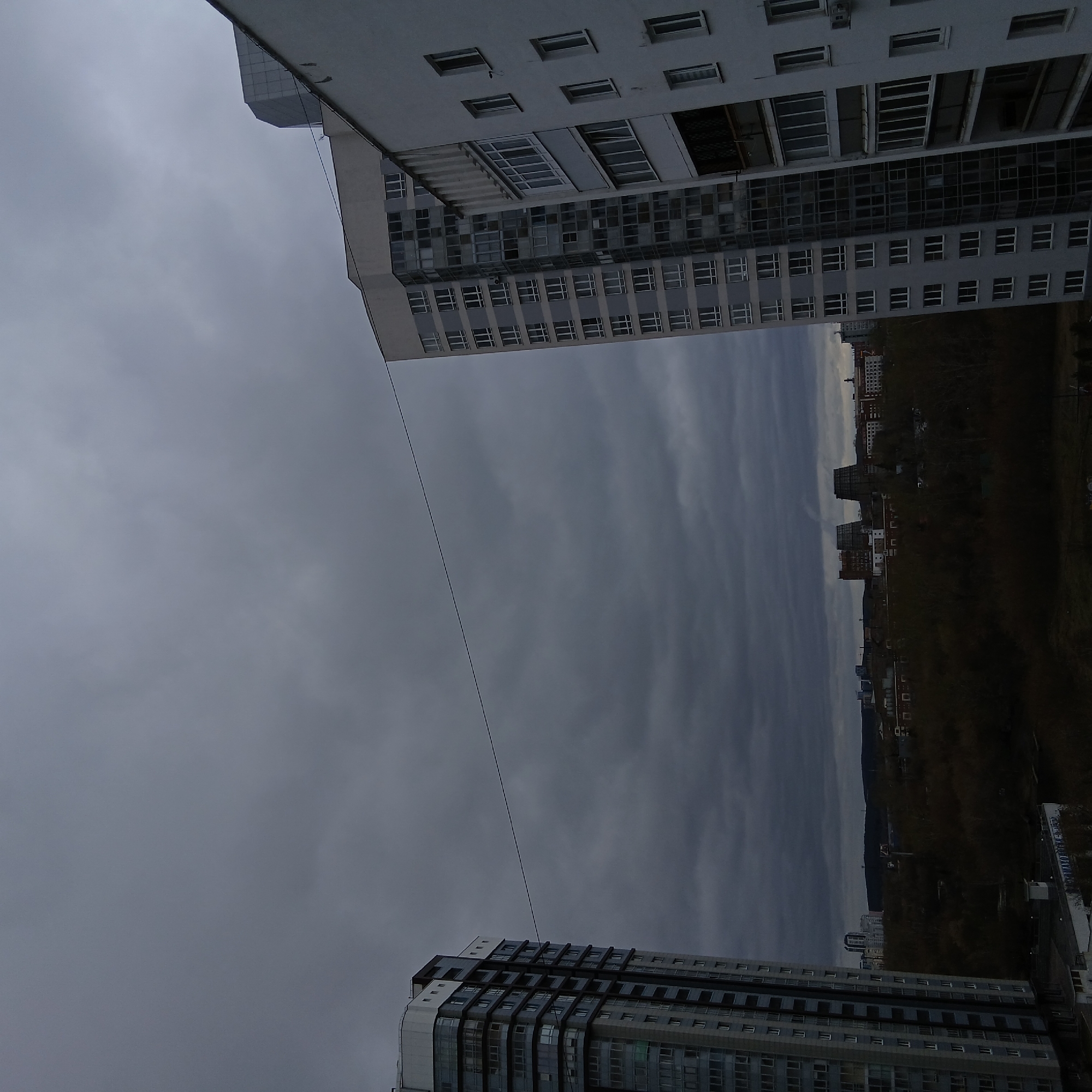 The image depicts a cityscape in Yekaterinburg, Russia under an overcast sky. The shot is taken from a lower vantage point, looking upwards and slightly tilted. Two high-rise buildings are prominent in the foreground: one on the upper left and one on the lower right. Both buildings feature multiple windows and are constructed with a modern aesthetic. A thin power line stretches diagonally across the frame, connecting the two buildings. In the background, a range of buildings and a densely wooded area are visible under the dark and cloudy sky. The overall tone of the image is somber due to the lack of sunlight and the gray color palette.