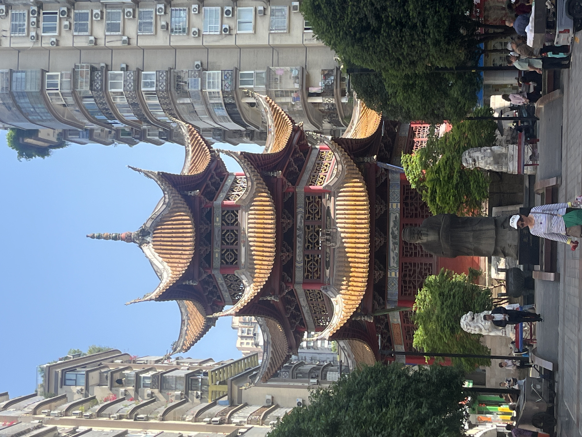 This image, taken in Jiangjin, China, features a prominent, ornate multi-tiered pagoda-style building at its center. The pagoda exhibits intricate traditional Chinese architecture with multiple upward-curving roofs covered in golden-brown tiles and decorated with detailed carvings and red accents.
Surrounding the pagoda are several tall, modern residential buildings, which serve as a stark urban backdrop. These buildings are light-colored and characterized by numerous windows, balconies, and visible air conditioning units. Lush green trees and bushes are interspersed around the pagoda and along a paved pathway to its right.
On the right side of the pagoda, a large, dark statue of a seated figure, possibly a historical personage, stands prominently. Further to the right, a white stone lion statue is visible near the pathway. Several people are scattered on the paved area, walking casually or standing, suggesting a public space like a park or square adjacent to the traditional structure.
The scene takes place on a bright, sunny day, indicated by the clear blue sky and strong illumination. The weather appears fair and pleasant. The overall impression is a blend of traditional Chinese culture, represented by the pagoda, set within a modern urban environment. No decipherable text is visible in the image.