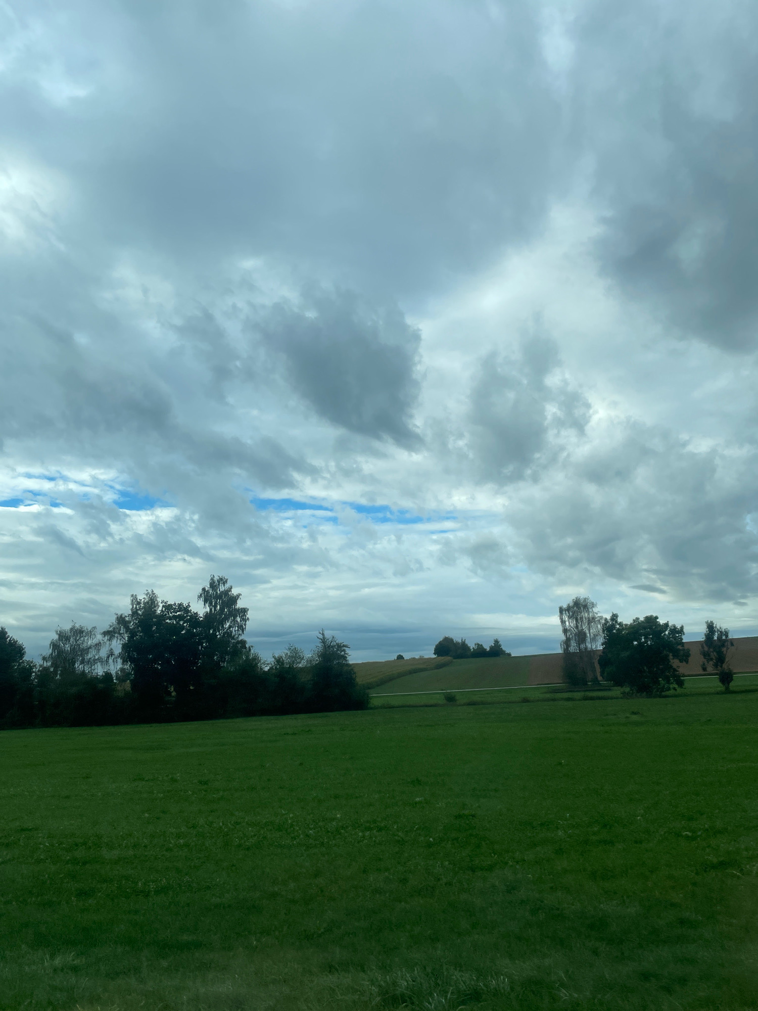 The image shows a wide view of a rural landscape under a cloudy sky. In the foreground, a lush green field stretches across the frame. Beyond the field, a line of trees separates it from a gently sloping hill. Further in the distance, a strip of land with cultivated fields can be seen, with some trees scattered throughout. The sky is filled with a dramatic display of grey and white clouds, with intermittent patches of blue sky visible. The overall scene evokes a sense of peacefulness and the vastness of nature.