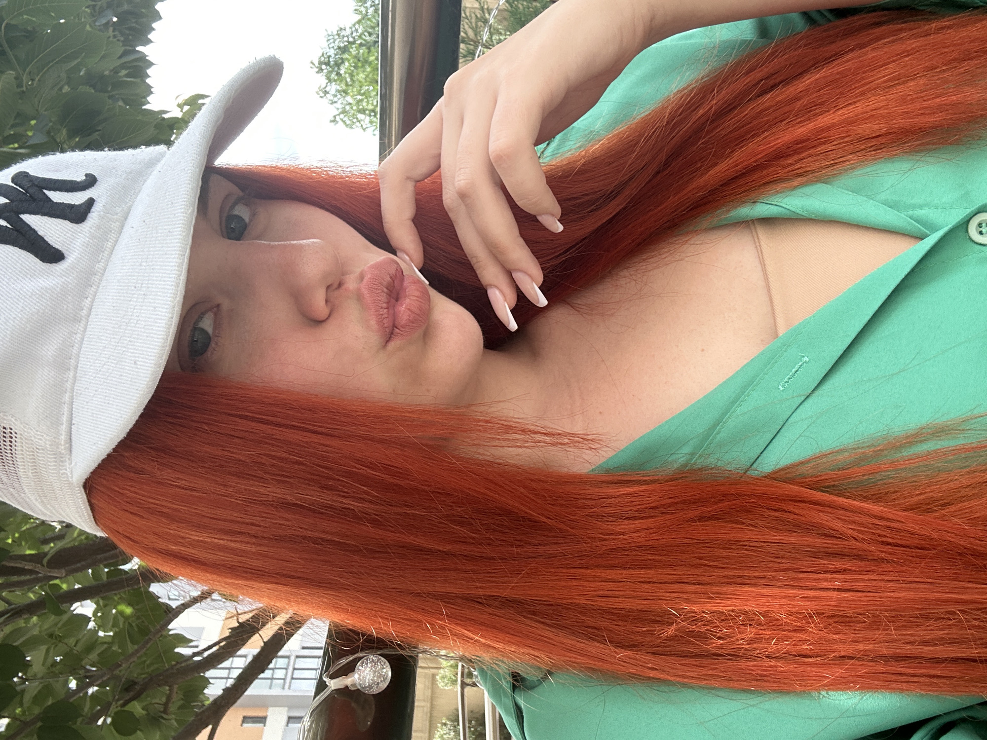 A woman with bright orange hair, wearing a white baseball cap with a black logo and a green shirt, poses for a selfie outdoors. She is puckering her lips and resting her hand on her chin, showing off her French manicured nails. In the background, green leaves from a tree are visible, along with a modern building. The lighting suggests it is daytime. The woman's attire and the presence of the building hint at an urban setting, possibly a park or a cafe area. The presence of the building in the background, consistent with modern architecture, supports the location in Lymanka, Ukraine, where such developments are common.