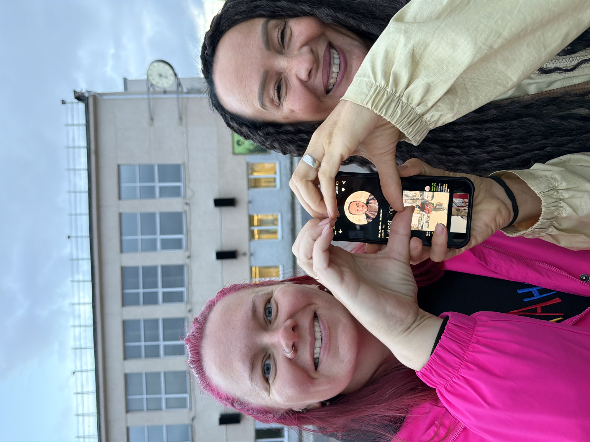 Two women are positioned in the foreground of an outdoor scene in Chaikovsky, Russia, against a light-colored building under an overcast sky. The woman on the left has bright pink hair, blue eyes, a nose ring, and is smiling, wearing a vivid pink jacket over a dark shirt with colorful embroidered letters "AH". The woman on the right has dark, braided hair, dark eyes, and is also smiling, wearing a light beige jacket and a silver ring on her left ring finger. Both women are holding a black smartphone with their hands, forming a heart shape around the phone's screen. The phone screen displays a profile picture of a man at the top, with text "Wahid_Rahman_Afghannai" and "RANKED #22", followed by "Latest Top". A smaller image of a street scene with a person on a bicycle is visible below this text. The background features a multi-story building with numerous rectangular windows, white frames, and multiple panes. An external round clock with a white face and dark numerals is visible on the right side of the building, and a railing structure is present on the rooftop. Several dark, rectangular fixtures are mounted on the building's facade. The natural lighting suggests daytime under cloudy conditions.