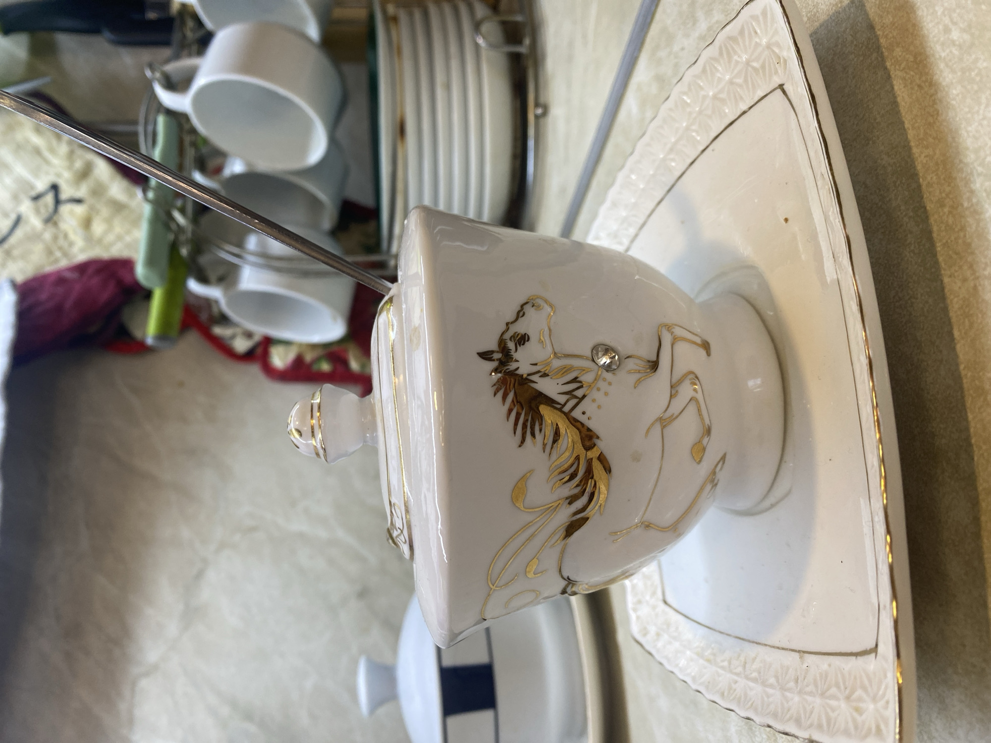 An ornate white porcelain sugar bowl with a lid, decorated with a gold horse design and a small crystal accent, rests on a matching square saucer with gold trim. A silver spoon is placed in the sugar bowl. In the background, white ceramic cups hang from a rack, and a stack of white plates with gold rims is visible. To the left, another white lidded dish with blue stripes is partially visible. The scene appears to be indoors, likely in a kitchen or dining area, on a patterned countertop. The location is identified as Novaya Usman', Russia.