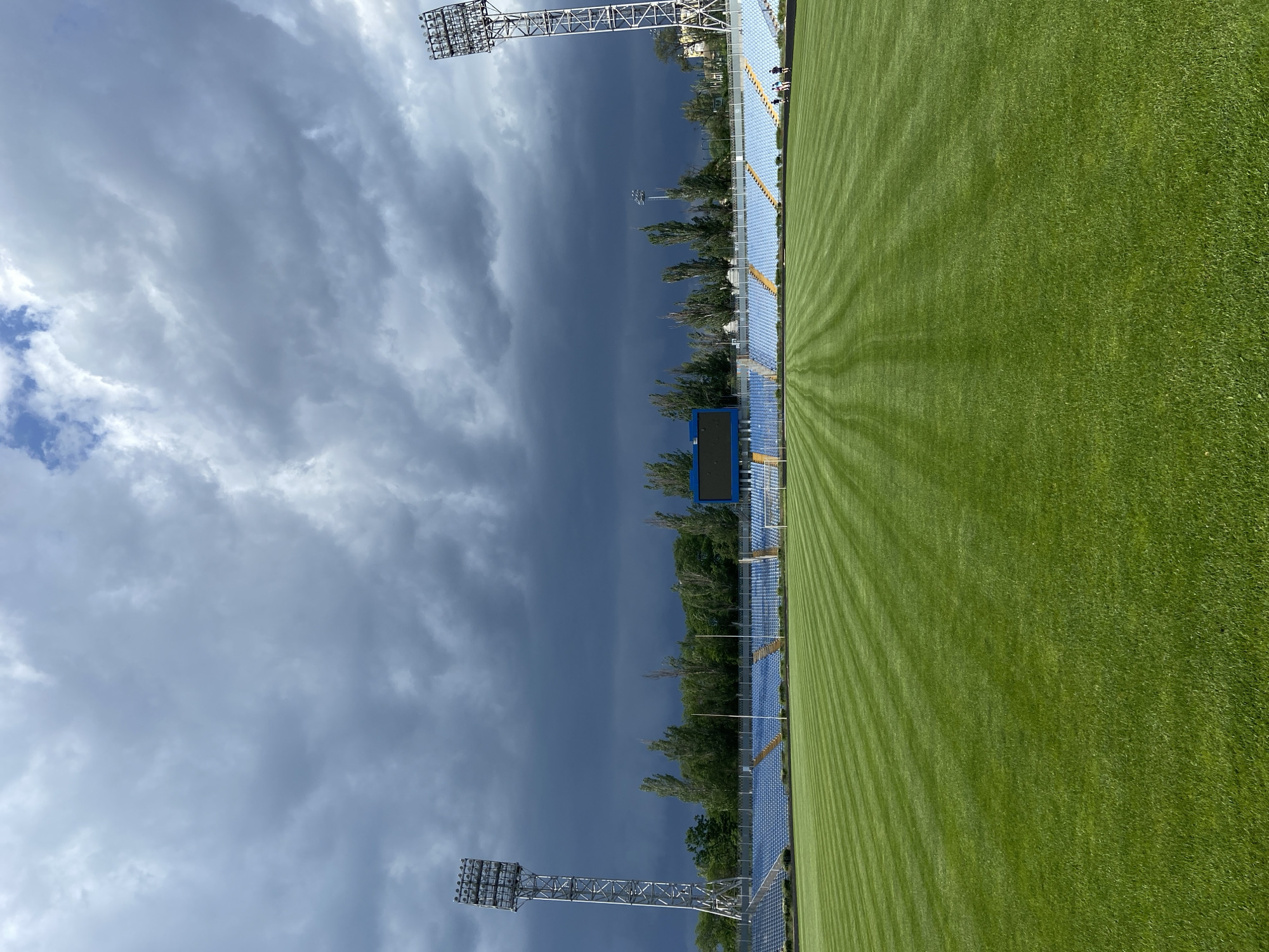 Here is a description of the image:
The image depicts a view of the "Metalurh Stadium" located in Kryvyi Rih, Ukraine. The perspective is taken from ground level along one side of the field. The field is green with visible mowing patterns. Blue seating lines one side of the field. Tall metal light towers stand at both ends of the visible section. A large, dark storm cloud looms in the sky. Some trees can be seen behind the seating.