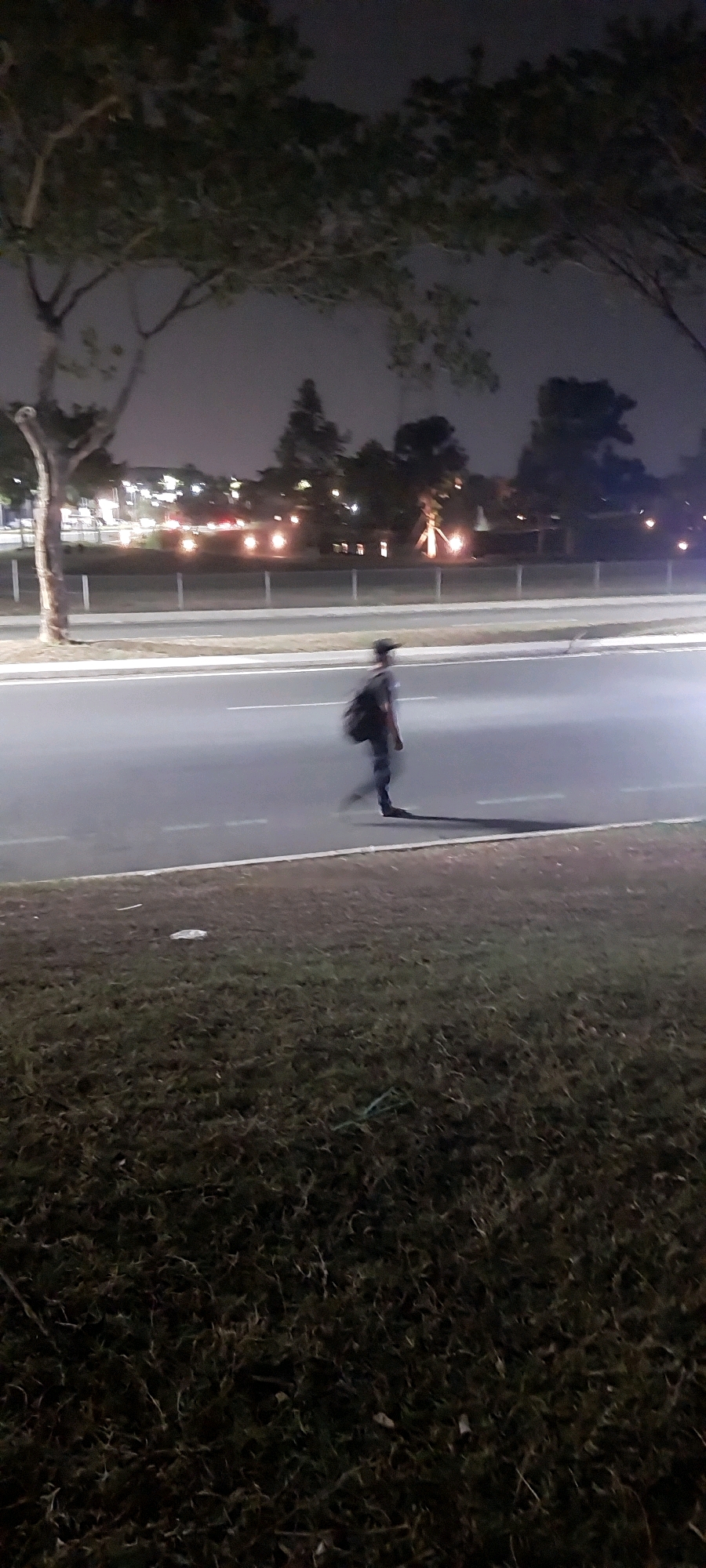A blurred person wearing a dark shirt, dark pants, and a baseball cap is walking across a paved road at night. The person is carrying a dark bag on their back. They are casting a long shadow on the road. The road has white lines marking lanes. To the left of the road, there is a grassy area with some sparse trees. In the background, there are streetlights and buildings, suggesting an urban environment. The scene is dimly lit, indicating it is nighttime, and the motion blur suggests the photo was taken with a slow shutter speed or while the camera was moving. The location is identified as a street in the city of Medan, Indonesia.