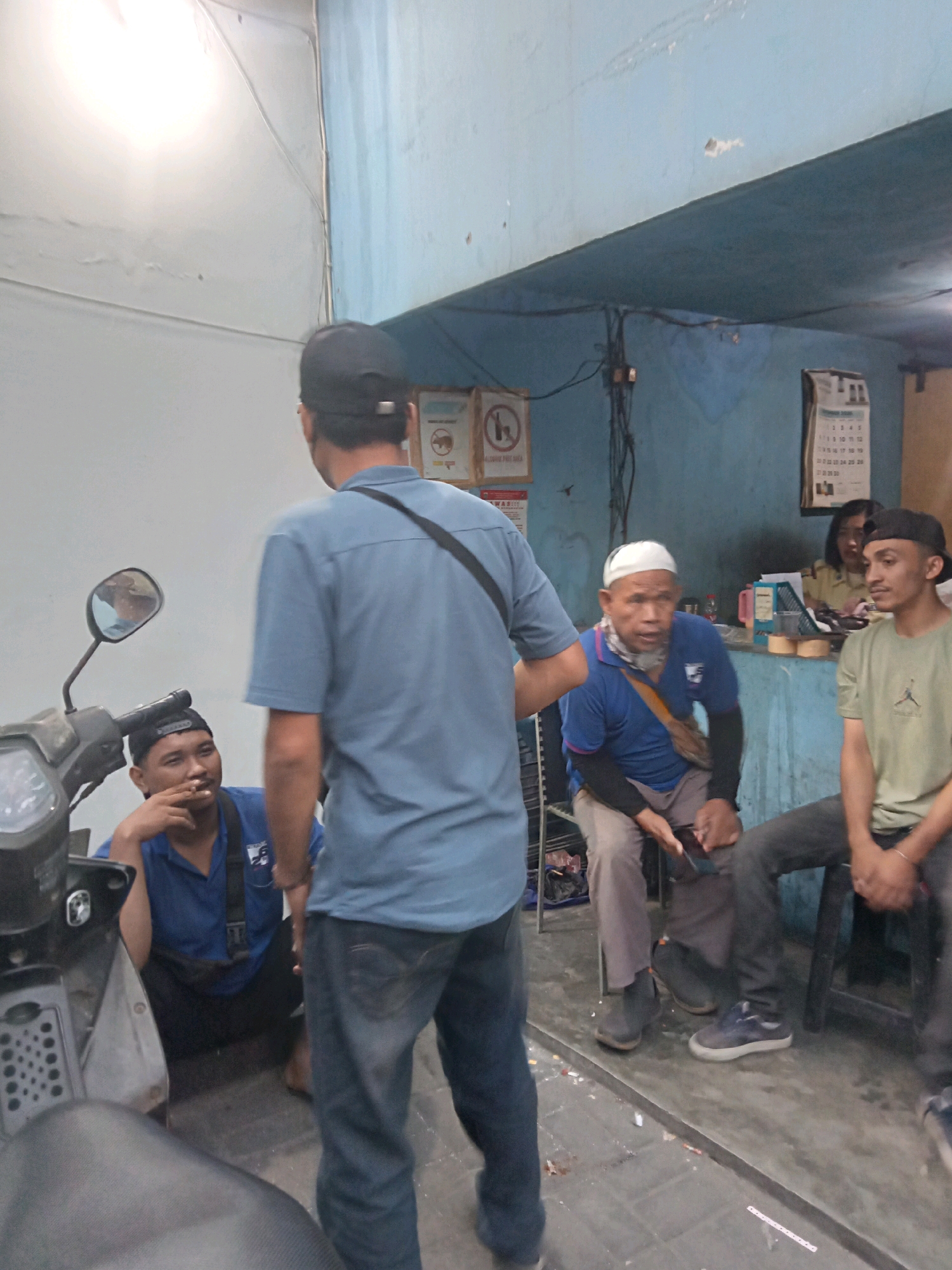 The image depicts an indoor scene with five individuals. In the foreground, a man wearing a blue shirt and a black baseball cap is standing and facing away from the camera, appearing to converse with another man. To his left, a man in a blue shirt and black baseball cap is sitting near a motorcycle, smoking a cigarette and looking towards the standing man. Further into the scene, a man in a blue shirt and white head covering is seated, holding a phone, and engaging in conversation. To his right, another man in a light green t-shirt is seated and looking forward. The environment appears to be a workshop or storage area, with blue walls, a light-colored ceiling, and a motorcycle in the left foreground. There are posters and a calendar visible on the back wall. The lighting is somewhat dim, with a bright light source in the upper left corner.