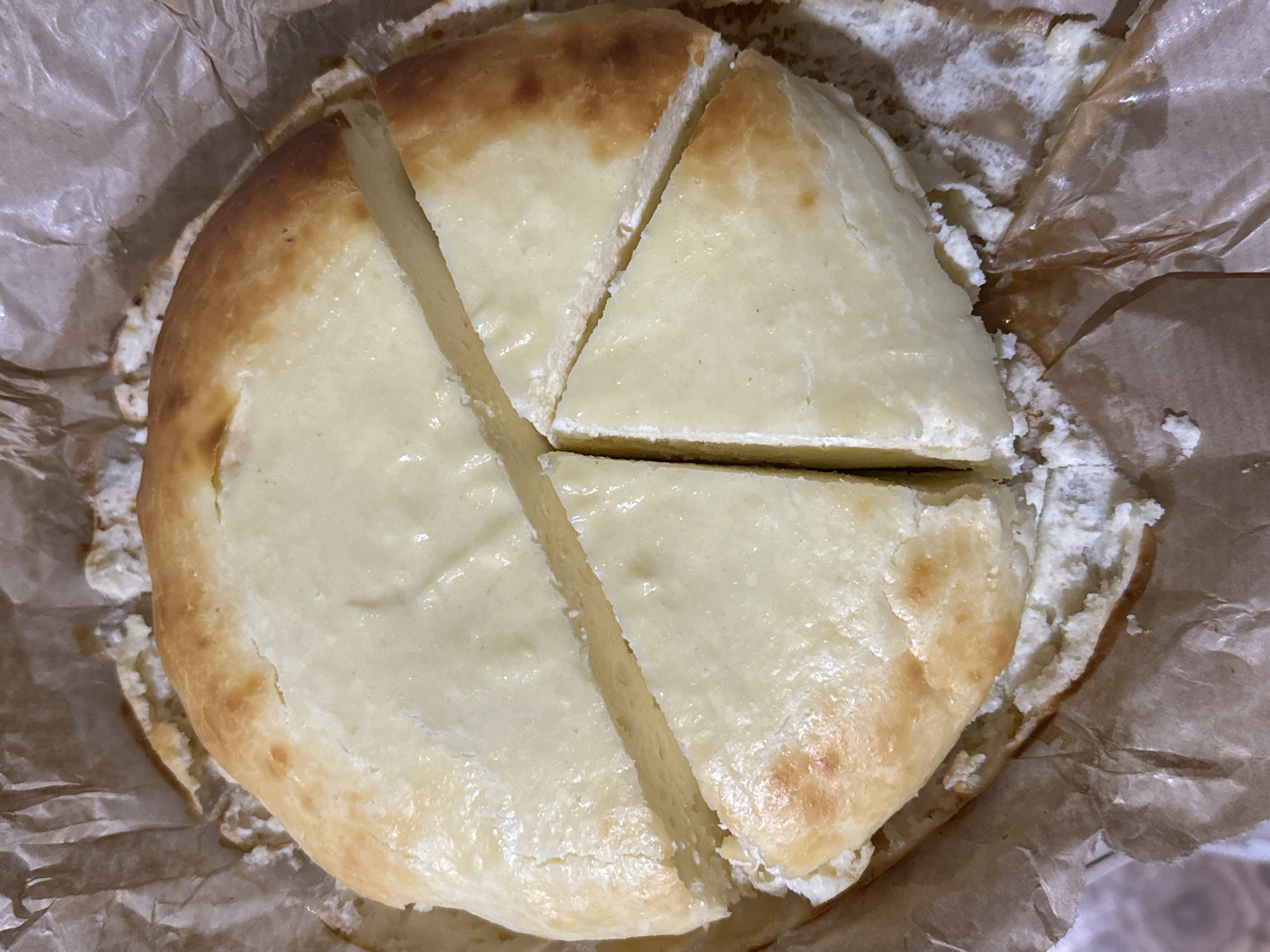 A close-up, overhead shot shows a round, baked dessert, possibly a cheesecake or sweet cheese tart, resting within crumpled light brown baking parchment. The dessert has been sliced into several irregular triangular pieces, with one slice partially separated from the main body.
The baked good features a smooth, pale cream-colored top surface, which appears slightly glossy and has minor textural variations, hinting at a creamy filling. The edges of the dessert are a light golden-brown, indicating a baked crust or edge, with some areas appearing slightly more browned than others. The cuts reveal a soft, white, somewhat crumbly texture inside, suggesting a cheese-based filling like cottage cheese or farmer's cheese.
The parchment paper surrounding the dessert is wrinkled and appears slightly moist or oily in some spots, with small crumbs visible, particularly where the dessert meets the paper. The overall impression is of a homemade or freshly baked item, ready to be served. No people or specific location cues beyond the food itself are visible in the image. There is no visible text. The lighting suggests an indoor setting.