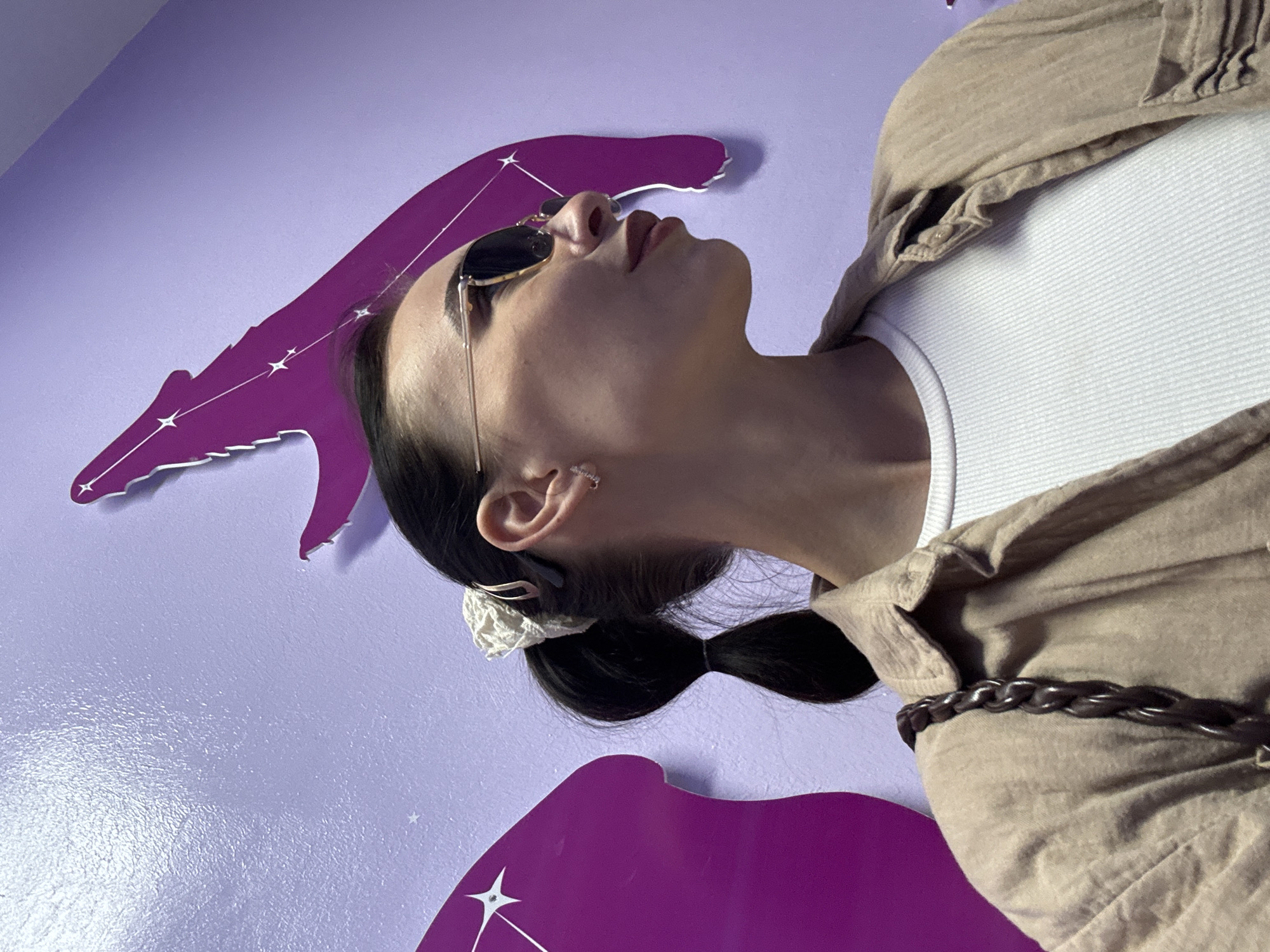 A low-angle, indoor shot shows a person with dark hair pulled back in a low ponytail secured with a white scrunchie. The person is wearing sunglasses and a beige shirt over a white tank top, and they are standing in front of a purple wall decorated with a purple constellation design. The person has an earring and is facing upwards with their head tilted back. The photograph was taken in Chaikovsky, Russia.
