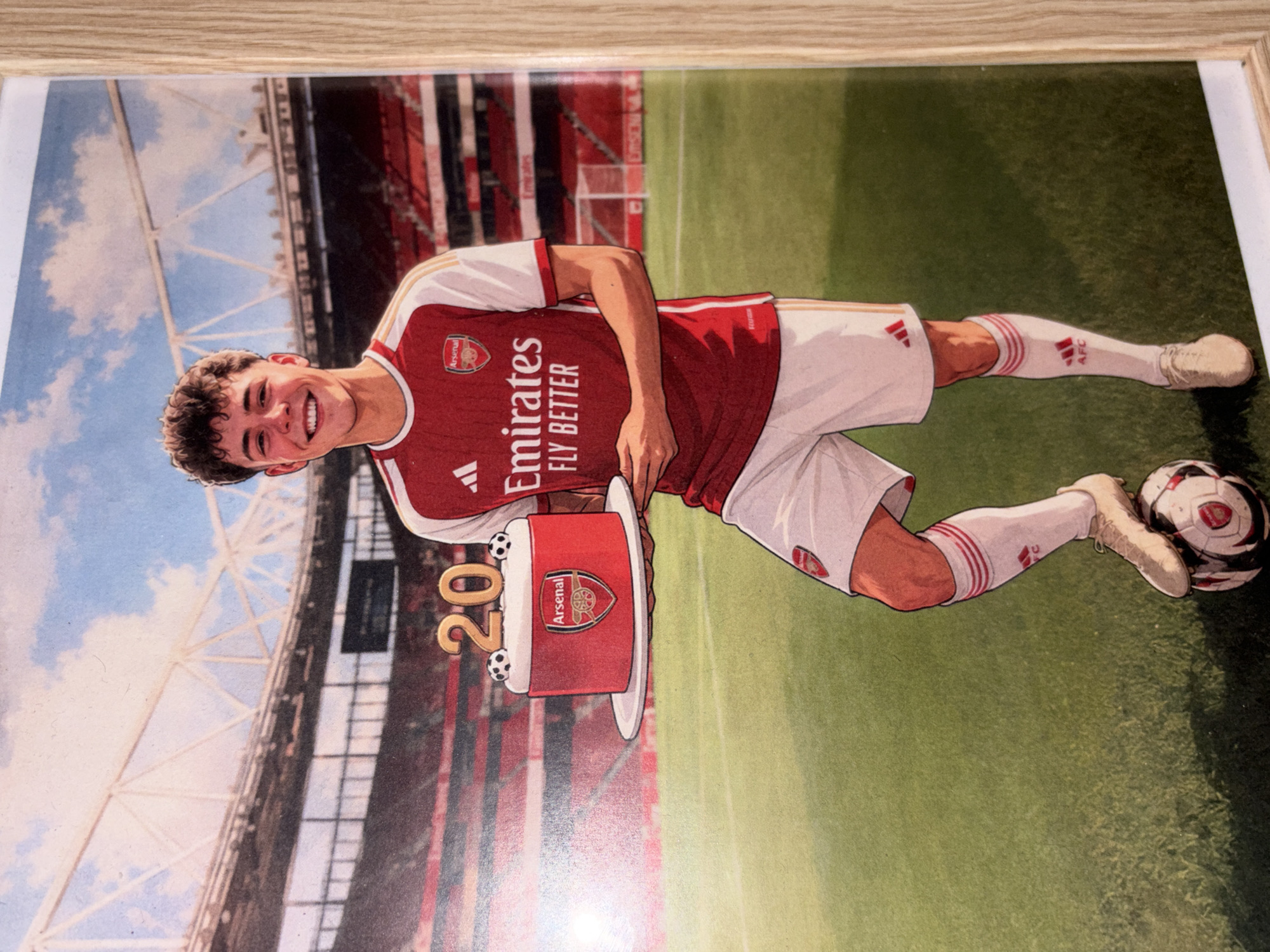 The image shows a framed cartoon drawing of a soccer player wearing an Arsenal jersey, white shorts, and matching socks and shoes. He is holding a cake that resembles a drum with the Arsenal emblem on it, adorned with the number "20". A soccer ball is on the ground next to his right foot. The background appears to be a soccer stadium with a cloudy sky above it. There is no information in the image indicating a location in Bezden, Bulgaria.