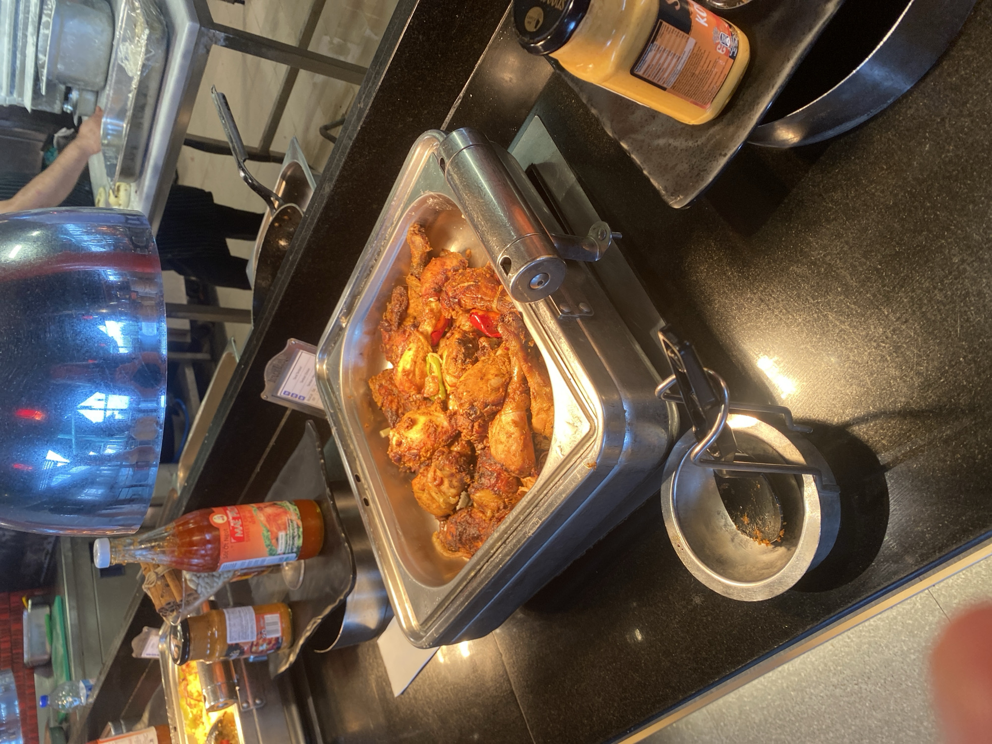 A buffet station displays a metal tray filled with roasted chicken pieces, some of which are garnished with red bell pepper slices. A silver serving utensil rests in a small metal bowl to the left of the tray. To the right of the chicken tray, a bottle of "Mai Tieu" sweet chili sauce stands upright. Further to the right, a yellow bottle of sauce, possibly mustard or curry, is positioned on a dark square plate. The buffet is set on a black countertop, with a metal warming lamp suspended overhead, casting a bright reflection. In the background, a person in dark clothing is partially visible near a stainless steel food preparation area. The scene appears to be indoors at a restaurant or hotel buffet, possibly in Aksu, Turkey, given the presence of local food items and typical buffet setup.