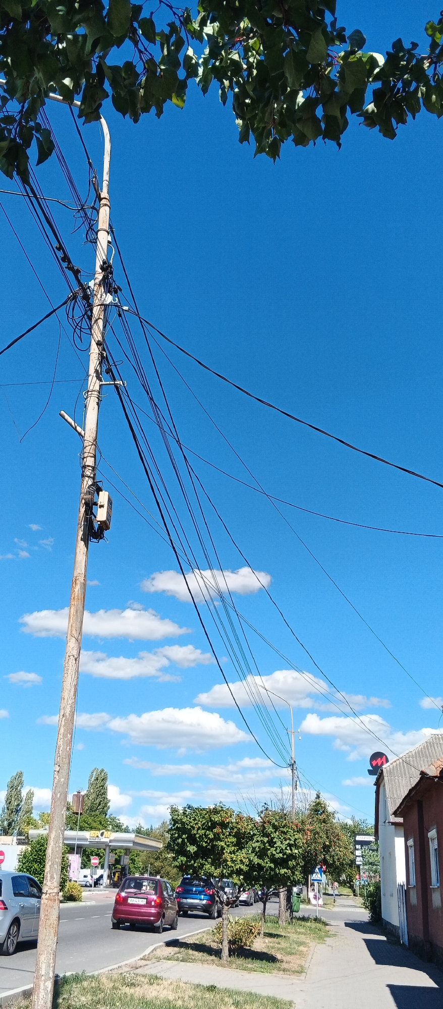 The image depicts a street scene in Mukachevo, Ukraine, on a sunny day with a clear blue sky and scattered white clouds. A wooden utility pole with a complex network of black wires dominates the left side of the frame, with a small white electrical box attached to it. In the foreground, on the right, is a sidewalk and the roofline of a building with a reddish-brown tile roof. A portion of a white and red sign with a stylized "M" logo is visible on the roof. The street below features several cars in motion and parked, including a silver hatchback on the left, a red hatchback, and a blue SUV in the center. Trees line the right side of the street and the sidewalk. In the background, a gas station with yellow and white signage is visible. Green foliage from overhead trees partially obscures the top of the image.