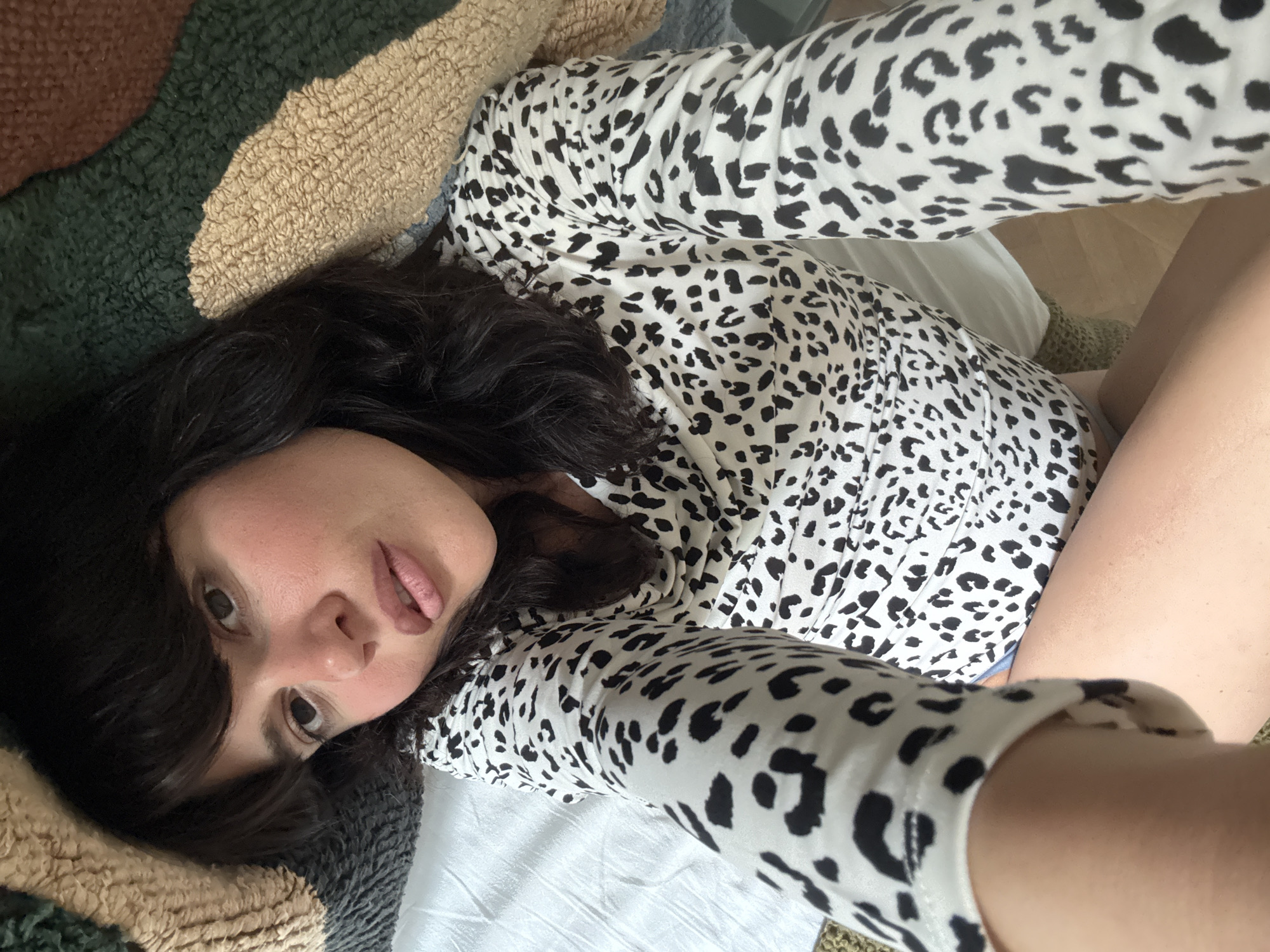 A woman is lying on a bed, taking a selfie. She is wearing a white top with black leopard spots. Her dark hair is spread around her head. The bed has white sheets, and a textured rug with brown, beige, and green stripes is visible. The location is unspecified.