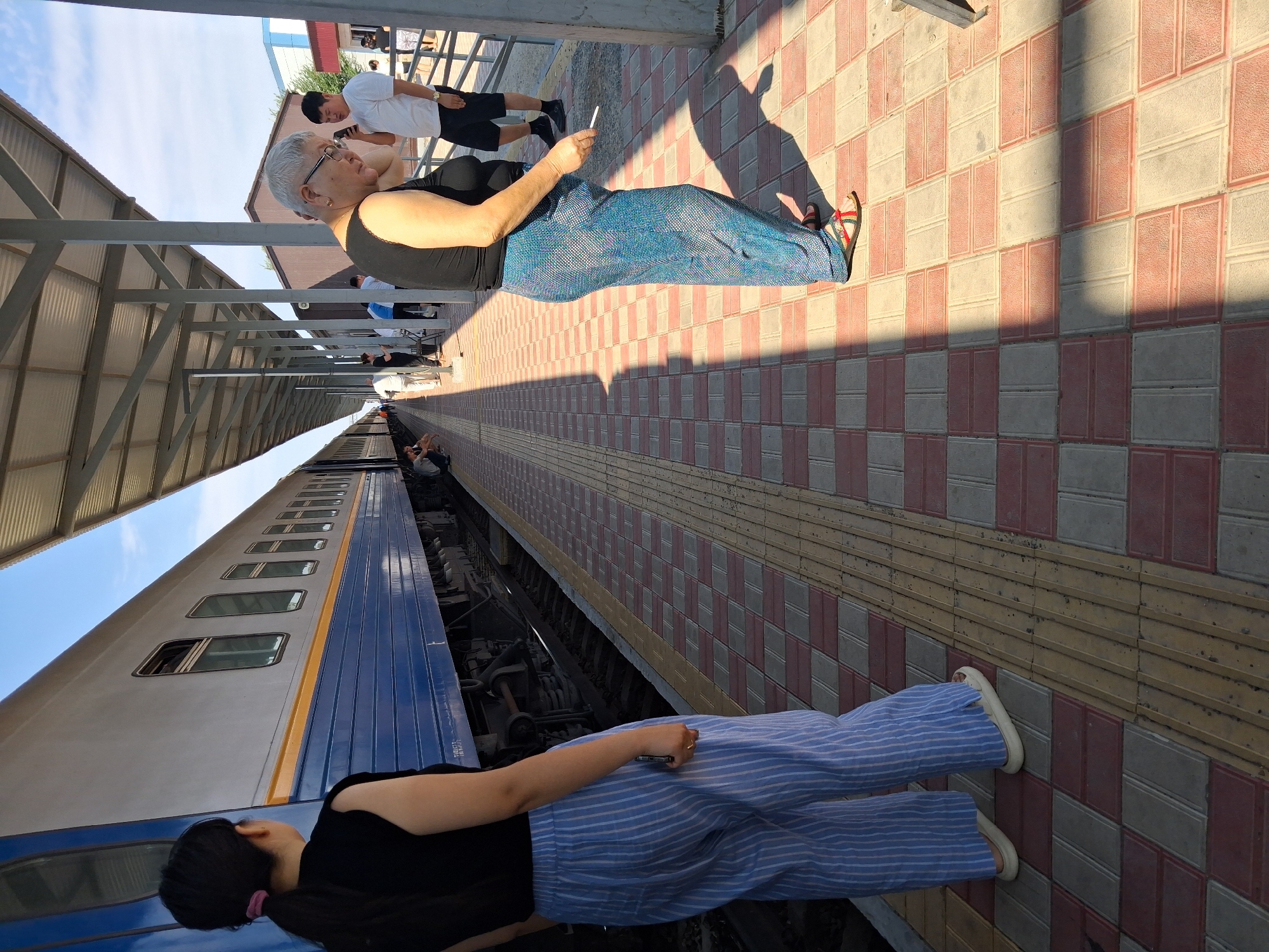 At a train station platform in Maqat, Kazakhstan, a young woman with dark hair pulled back in a ponytail stands in the foreground, wearing a black sleeveless top and light blue and white striped wide-leg pants. She faces away from the camera, looking towards a parked train. To her right, an older woman with short gray hair and glasses wears a black tank top and blue patterned wide-leg pants. She holds a white cigarette in her right hand and looks down, seemingly at her phone, which is in her left hand. Behind her, a young man in a white t-shirt and black shorts stands, also looking at his phone. The platform is paved with alternating red and light-colored rectangular tiles, with a textured yellow tactile paving strip along the edge. A covered walkway with metal supports runs along the platform, casting long shadows. The train, with blue and yellow detailing, is visible on the tracks. Other people are visible in the background, some sitting on the platform and others walking. The sky is clear and blue.