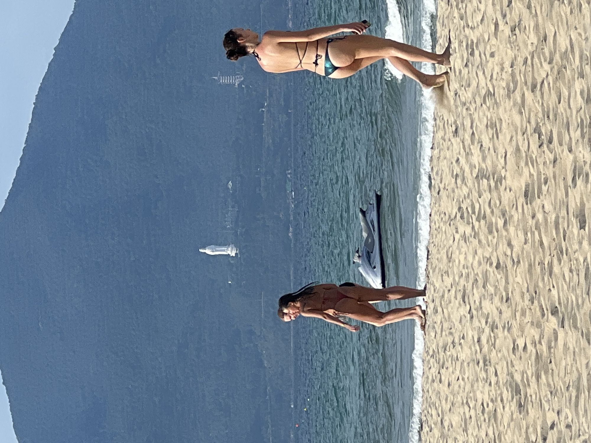 This is a medium shot of a beach scene in Da Nang, Vietnam, with a large, verdant mountain in the background. A white statue of a deity, likely the Lady Buddha, is visible on the mountainside. Two women are walking on the beach in bikinis. The woman on the right is walking away from the camera, and the woman on the left is walking toward the camera. A jet ski is partially visible in the water near the women. The ocean is blue and calm, with small waves lapping at the shore. The sand is light brown and appears to be dry. The sky is clear and blue. The overall impression is a sunny day at the beach.