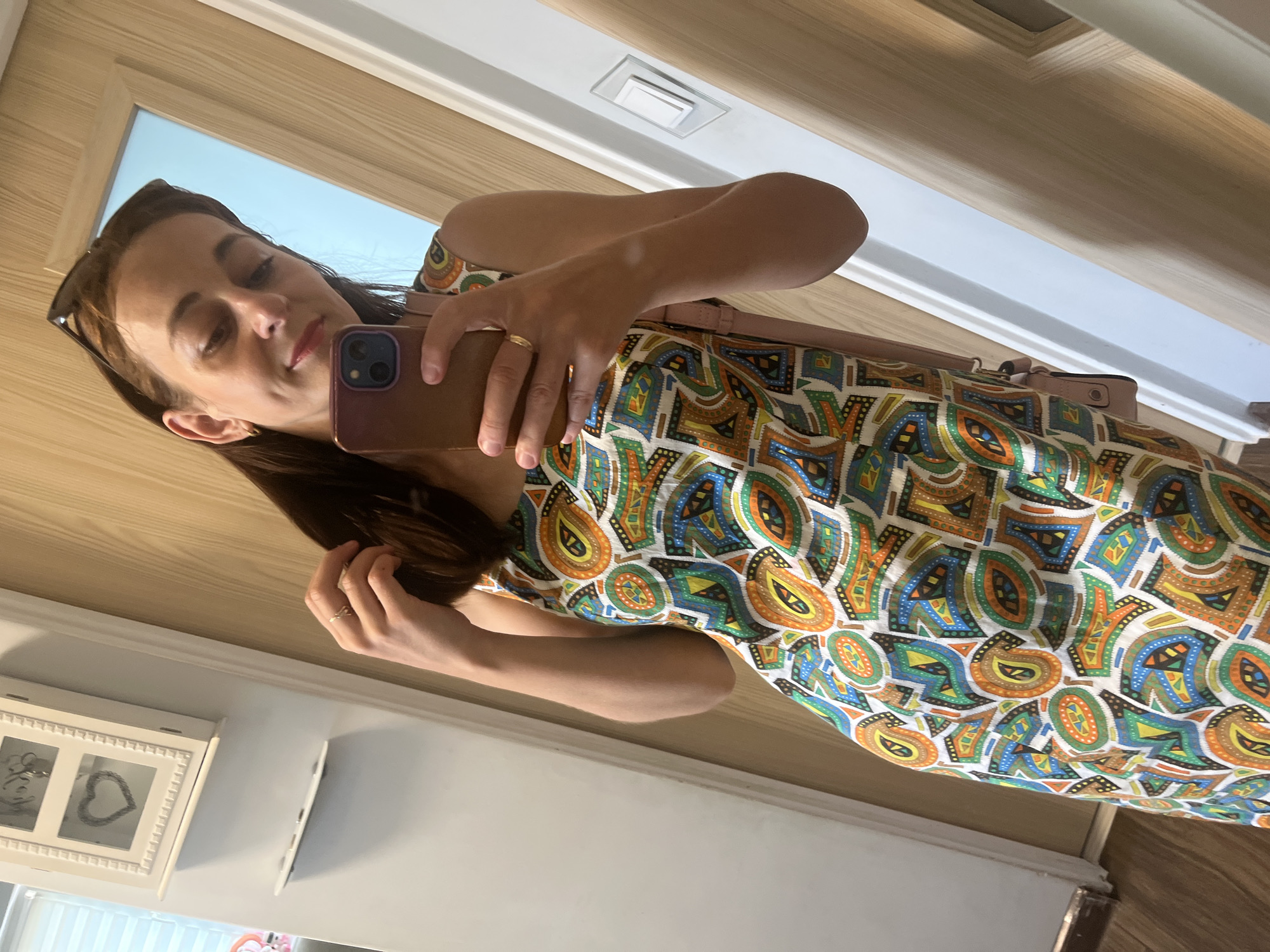 A woman, whose face is partially obscured by a mobile phone, is taking a mirror selfie in a hallway. She is wearing a sleeveless dress with a colorful, geometric pattern. Her right hand is raised, holding the phone, and her left hand is touching her hair. A small, light pink purse is slung over her left shoulder. The background shows a white wall on the left, a wooden door and frame on the right, and a wooden floor. A decorative white wall-mounted cabinet with a heart-shaped cutout is visible on the left wall. A light switch is mounted on the right wall. The overall environment appears to be indoors, likely a residential setting.