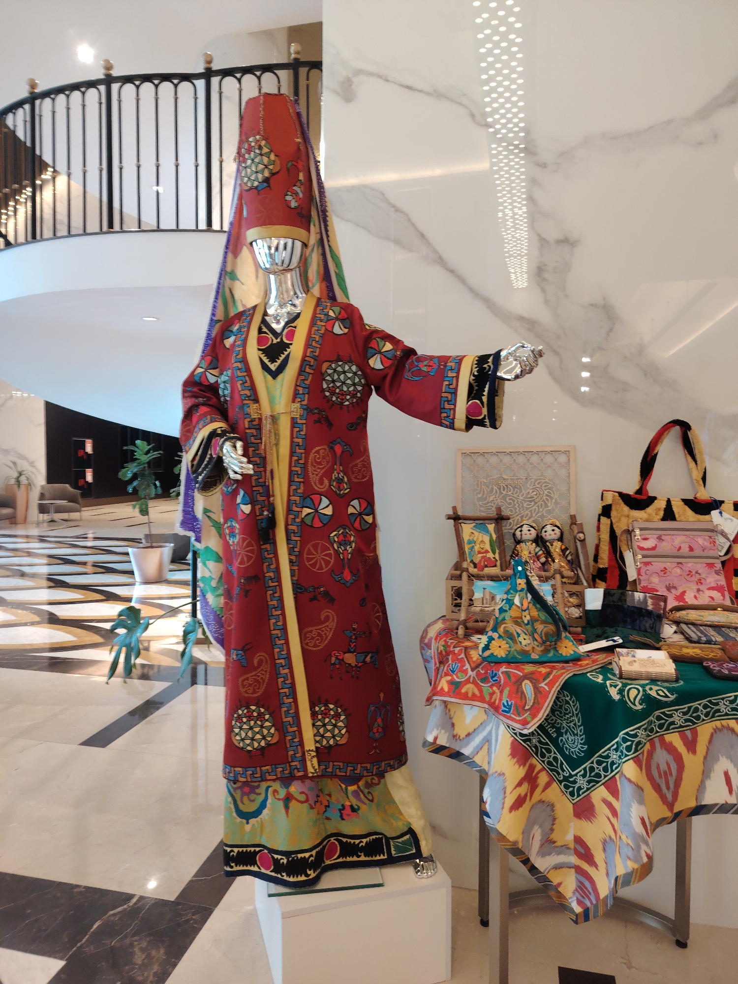 In Konigil, Uzbekistan, a mannequin stands in a well-lit, modern interior, likely a reception area or display space. The mannequin is dressed in an elaborate, red, traditional robe adorned with intricate patterns, including circular motifs and stylized figures. A tall, conical red headdress, also decorated, is worn by the mannequin, with a colorful fabric draped behind it.
To the right of the mannequin, a table is laden with various handcrafted items. These include two small dolls, several embroidered pouches or coin purses in shades of pink and beige, and a large, colorful tote bag with red, black, and yellow geometric patterns. A green textile with white embroidered patterns is draped over the table, as are other smaller decorative cloths with vibrant, patterned borders, such as yellow and red ikat designs. A wooden frame with an openwork pattern rests on the table behind the dolls.
The environment features a polished, checkered marble floor with a distinctive geometric pattern in black and off-white. In the background, a black metal railing with gold finials curves around a staircase. The walls are finished with white marble slabs. The overall impression is one of a cultural display or a boutique showcasing traditional Uzbek crafts within a contemporary setting.