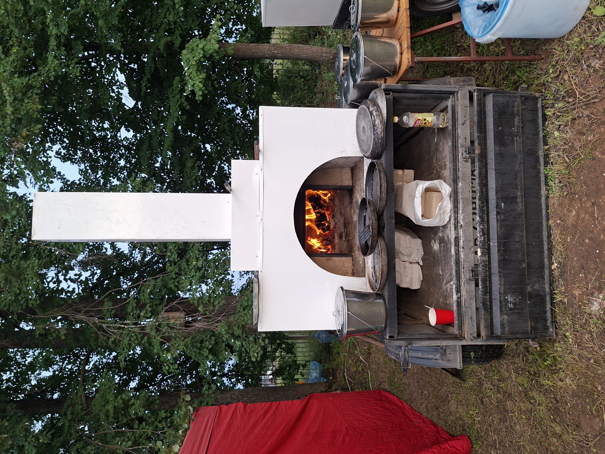 The image depicts an outdoor scene, possibly in Старые Быги, Russia, featuring a portable wood-fired pizza oven mounted on a trailer. The oven is white with a black arched opening where flames are visible, indicating it is in use. Above the oven, a white chimney extends upwards. A red tent is partially visible on the left side of the frame, and lush green trees with dense foliage form the background.
In front of the oven, on a black shelf, are several cooking implements, including frying pans and a metal bucket with a lid. To the right of the oven, a wooden table holds more metal buckets. Below the shelf, the trailer's storage compartment is visible, containing a red plastic cup with a stirrer, white bags that appear to hold firewood, and a bottle of cooking oil. A blue trash bin with a blue bag liner is situated on the ground to the right of the wooden table. The ground is covered with grass and dirt. The lighting suggests it is either early morning or late evening. There are no people visible in the image.
