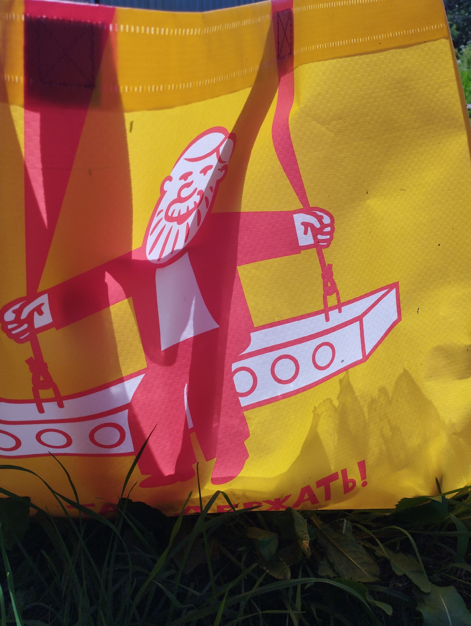 A yellow fabric shopping bag with a red graphic illustration of a man is resting on the ground amidst green grass and leaves. The illustration depicts a man with a beard and a red jacket, holding a pole connected to a white object with red circles. A portion of red text, likely part of a Russian phrase, is visible on the bottom right of the bag, reading "РЖАТЬ!". The bag is illuminated by sunlight, casting shadows on its surface. The background shows some blurry greenery, suggesting an outdoor setting in the city of Shiryaevo (Ilinsky), Russia.