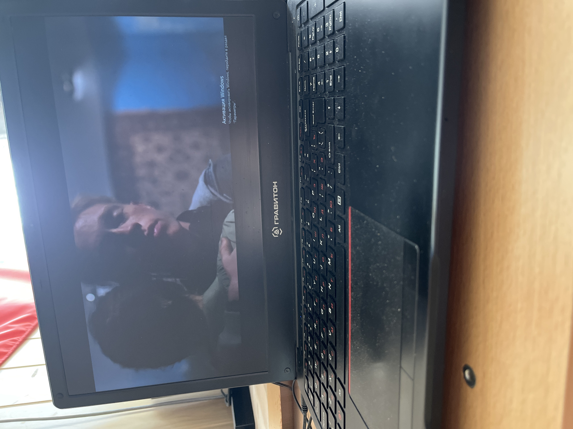 Here is a detailed description of the image:
**Scene Overview:**
The image shows a laptop, seemingly indoors. The laptop screen displays what appears to be a movie or video paused, with a partial view of a person on screen. The laptop is unactivated Windows, according to text in Russian.
**Objects:**
* **Laptop:** The primary object in the frame is a black laptop. The brand "ГРАВИТОН" is visible on the screen bezel. The keyboard has a red accent on the right edge. There is dust present on the keyboard and surface of the laptop.
* **Screen Display:** The laptop's screen shows a still from a movie or video, with the figure of a person visible.
* **Text on Screen:** "Активация Windows" (Windows Activation) is displayed on the screen in Russian.
* **Table/Surface:** The laptop rests on a wooden table.
**People:**
* **Person on Screen:** A face is partially visible on the laptop screen.
**Actions:**
* **Paused Video:** The laptop screen indicates a paused video.
**Environment:**
* **Indoor:** The image suggests an indoor setting based on the lighting and visible wooden surface.
* **Russia:** The image may be located in the city of Chaikovsky, Russia given that the language on the screen is Russian and that the brand "ГРАВИТОН" is known to have a presence in Russia.
