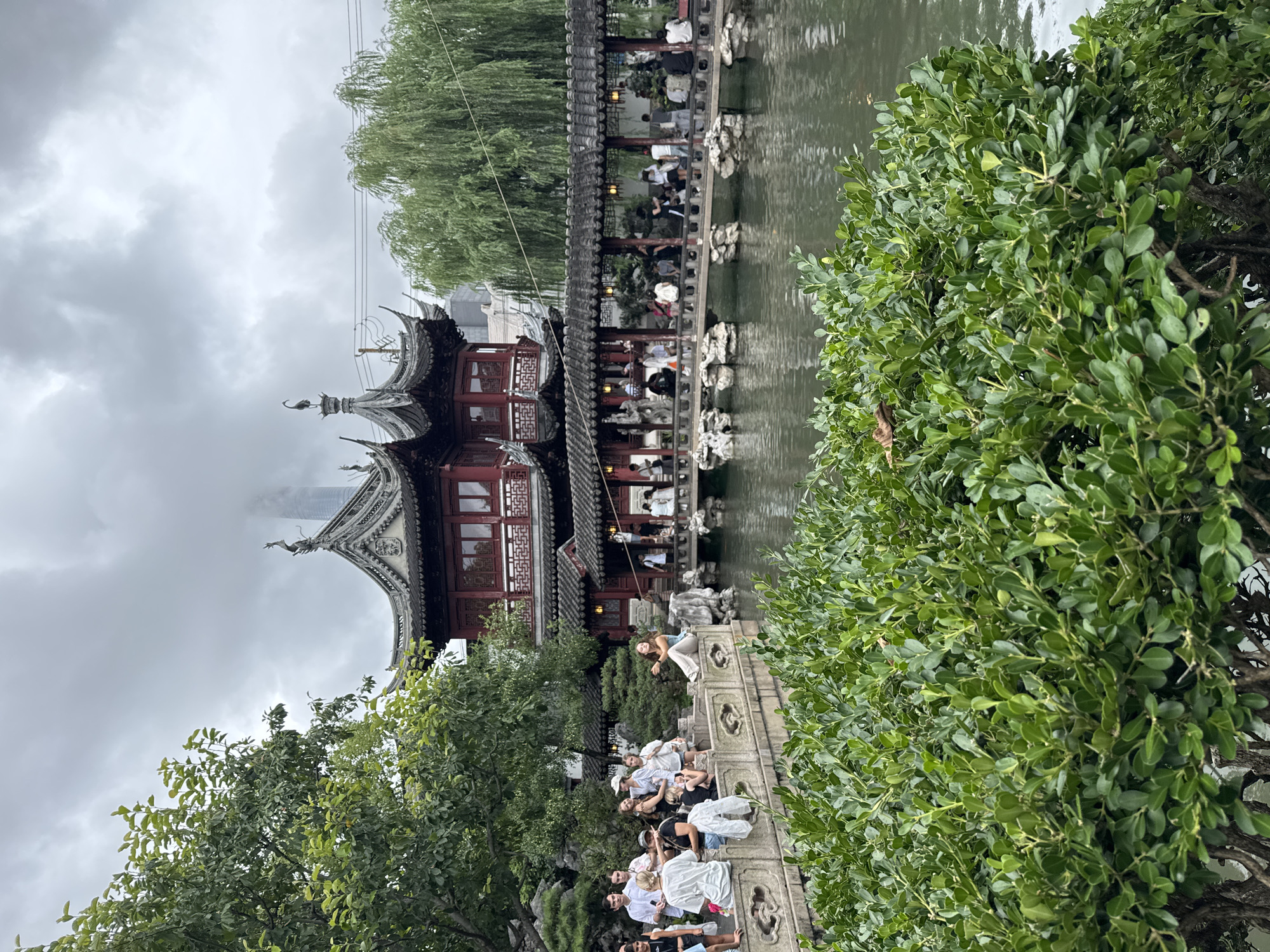 This is a photograph of the Yu Garden in Shanghai, China, on a cloudy day. The foreground is dominated by lush green bushes. In the middle ground, a body of water reflects the overcast sky and the surrounding architecture. A stone bridge with intricate carvings spans the water, connecting a walkway to a traditional Chinese pavilion. The pavilion features multiple tiers with ornate, dark tiled roofs and red accents. People are visible on the bridge, the walkway, and a stone railing on the left, observing the garden and the architecture. Some are sitting, others are standing and talking. Trees, including a large leafy tree on the left and a weeping willow on the right, frame the scene. In the background, modern high-rise buildings are visible through the cloudy sky, providing a stark contrast to the historical garden. The overall atmosphere is serene, with a sense of historical tranquility juxtaposed with the bustling modern city.