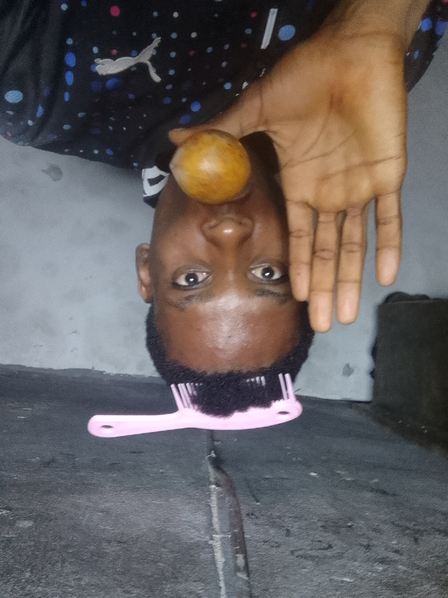 A man lies inverted on a gray concrete surface. He wears a dark, patterned shirt with a white Puma logo. An orange fruit rests on his upper lip. His right hand is open beside his face. A pink comb is positioned on his hair, facing the bottom of the frame. The location is Unknown, Unknown.