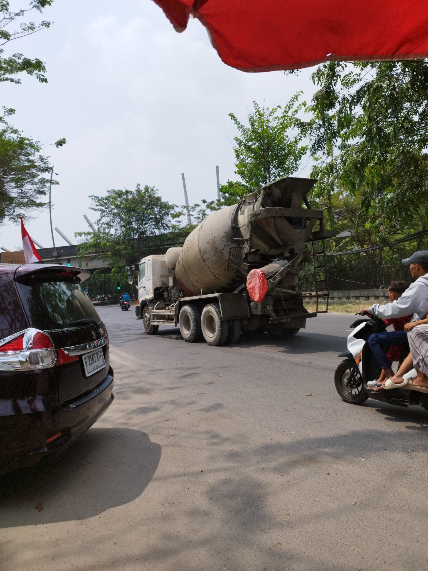 The image captures a street scene in Bekasi, Indonesia, characterized by a cloudy sky and lush green trees lining the road. In the foreground, a dark-colored SUV is parked on the left side of the road, displaying a license plate that reads "B 2365 KYP". A small Indonesian flag is affixed to the rear of the SUV. To the right, a cement mixer truck dominates the mid-ground, appearing dusty and well-used. On the right side of the road, a scooter carries three people: a driver wearing a grey hoodie and a baseball cap, a child in a red shirt, and a person in the back wearing a patterned skirt. The scooter and its passengers are captured in motion, moving away from the camera. In the background, a pedestrian bridge spans across the road, with overhead wires also visible. The overall environment suggests a typical urban or suburban setting in Bekasi.