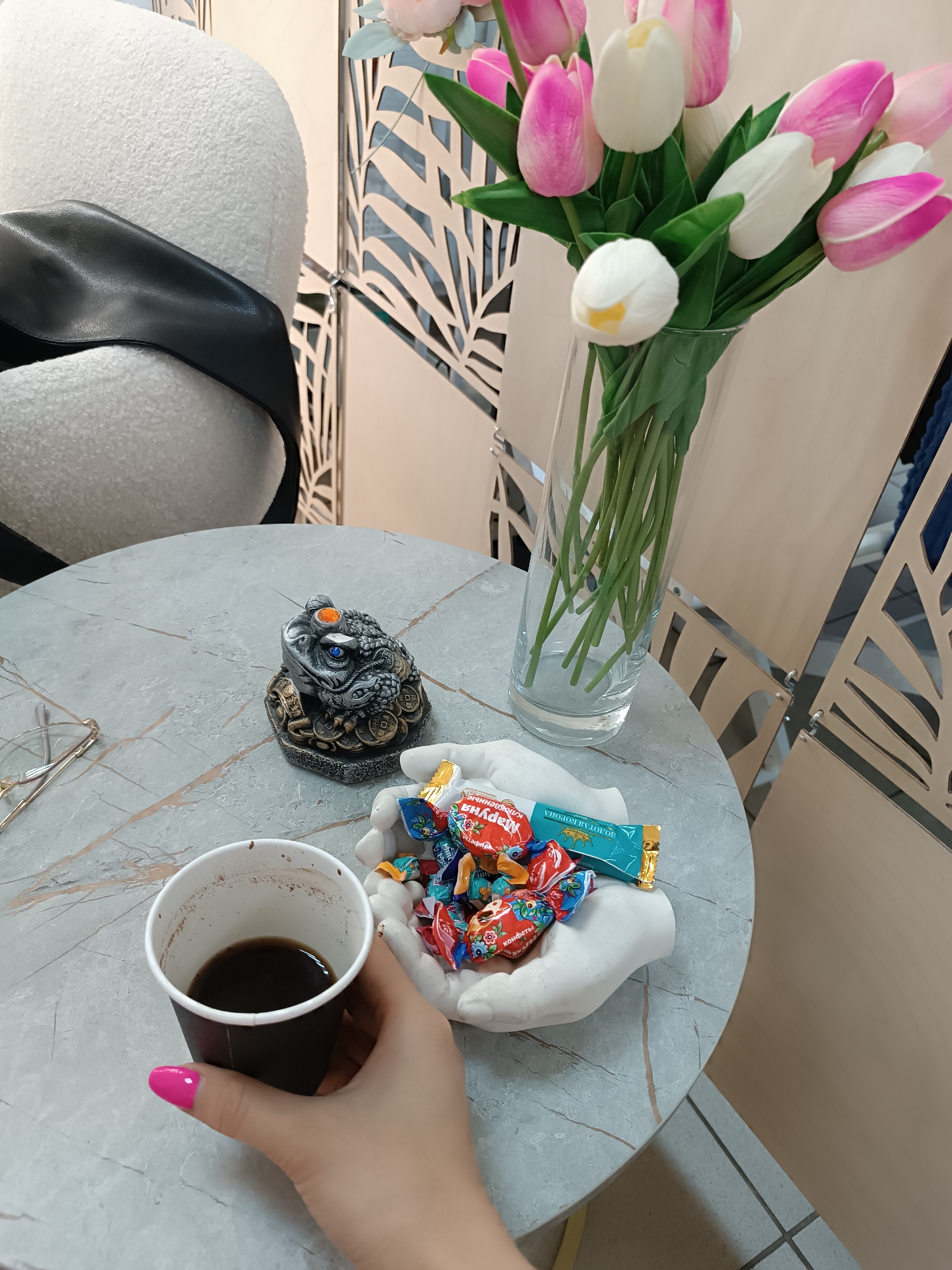 A high-angle shot captures a tabletop scene, likely indoors, in an unspecified city. A person holds a black paper cup containing a dark liquid, presumably coffee, with a manicured hand featuring bright pink nail polish. On the table, there is a pair of spectacles, a stone frog figurine on a bed of coins, and a pair of white ceramic hands holding a colorful assortment of Russian wrapped candies. A clear glass vase filled with artificial tulips in shades of pink and white adds a touch of floral decor. A white chair with a black strap is in the background. The table has a marble-like finish. A decorative metal screen with a leaf pattern is behind the vase. The lighting appears to be soft and ambient.
