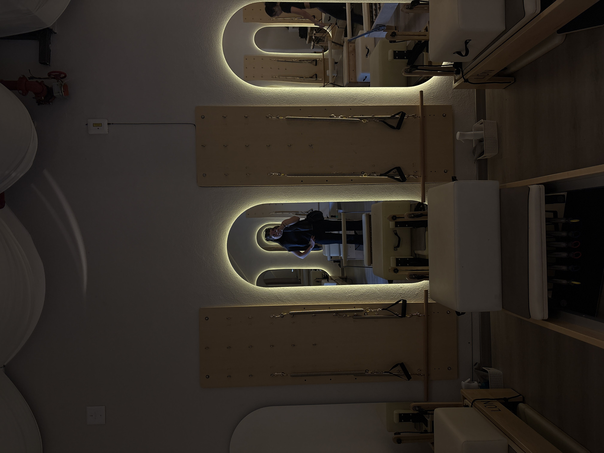 The image depicts the interior of a Pilates studio, as reflected in a series of backlit, rounded rectangular wall mirrors. Each mirror is mounted above a wooden Pilates wall unit with attached springs and handles. In one of the mirrors, a woman wearing dark clothing is visible. The walls are painted in a light color. Pilates equipment and accessories are visible on the right side of the image, including reformer machines. The location is likely in Mexico City, Mexico.