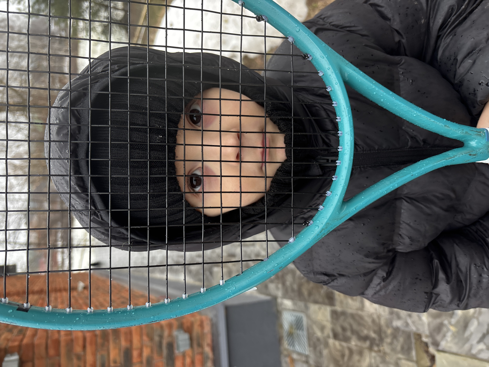 A young child, wearing a black knit hat and a black puffy jacket, is holding a turquoise tennis racket up to their face, with the racket's strings creating a grid-like pattern across their face. The child's face is visible through the racket strings, showing their eyes, nose, and mouth. Water droplets are visible on the tennis racket and the jacket, suggesting it has recently rained.
The background is blurred and indistinct, but the presence of brickwork at the bottom left corner of the frame implies that the location is in an urban area, most likely in the city of Chaikovsky, Russia. Additionally, what appears to be a paved pathway is also visible on the right side of the frame.