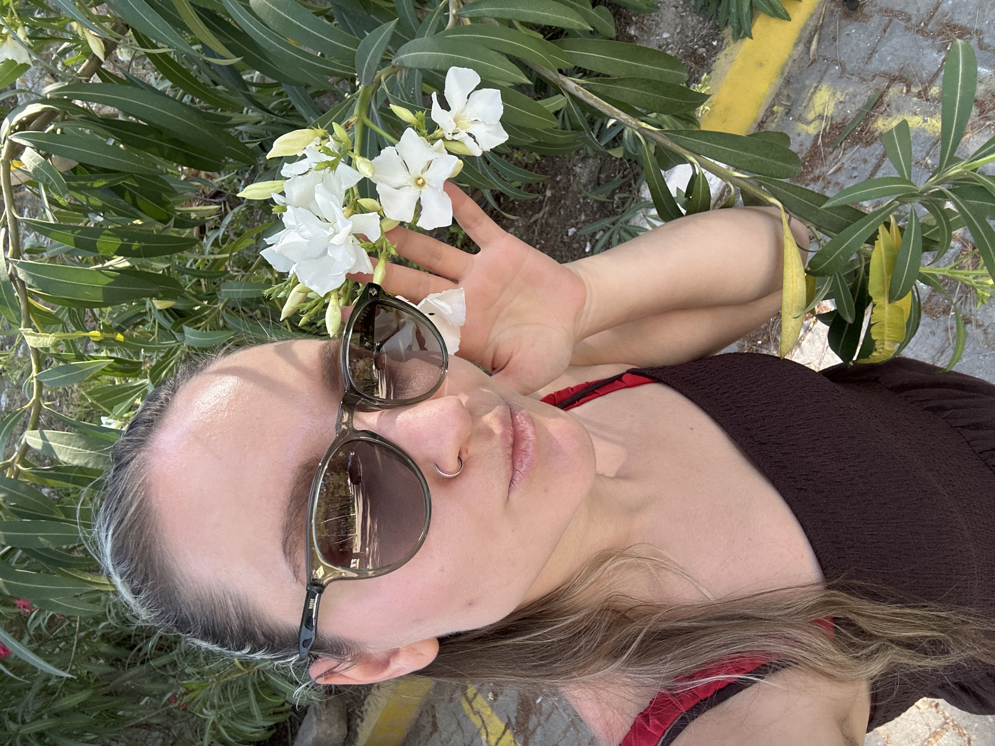 The image shows a woman lying down, her head near white flowers and green leaves of an oleander bush. She is wearing sunglasses, a brown top with red straps visible, and a nose ring. The ground is visible in the background, showing pavement with yellow markings. Given the presence of oleander bushes and pavement, the location is likely an urban or semi-urban setting. Considering the prompt, the location is Fethiye, Turkey. There are no evident actions or interactions.