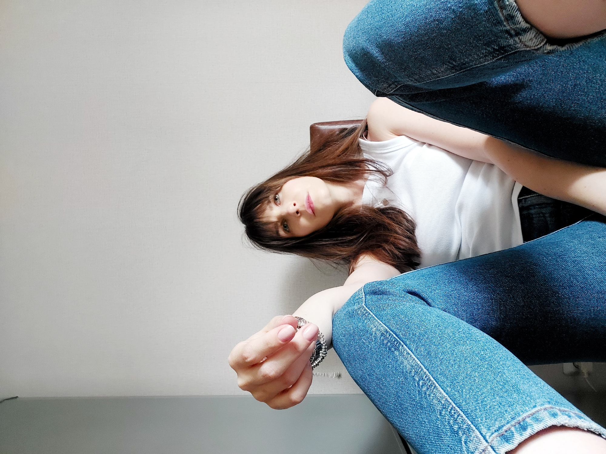An image, rotated ninety degrees counter-clockwise, depicts a young woman from a low-angle perspective, appearing to recline or lie down. Her head is towards the left of the frame, with her gaze directed upwards towards the camera. She has long, dark brown hair with bangs, fair skin, and light-colored eyes, with a calm and slightly engaging expression. Her lips are subtly parted.
She is dressed in casual attire, consisting of a white ribbed tank top and blue denim jeans. The jeans show some distressed elements, particularly a torn hem on one leg. On her left wrist, she wears a silver-toned bracelet. Behind her head and shoulder, a portion of a brown, possibly leather or faux leather, chair armrest is visible.
The background is simple, featuring a plain, light grey or off-white wall in the upper left. Below this, a darker grey, possibly metallic, surface extends across the lower part of the frame, likely a table or floor. The setting appears to be indoors, under ambient lighting. There are no specific visual cues within the image to indicate time of day, weather, or a precise location. No text is visible.