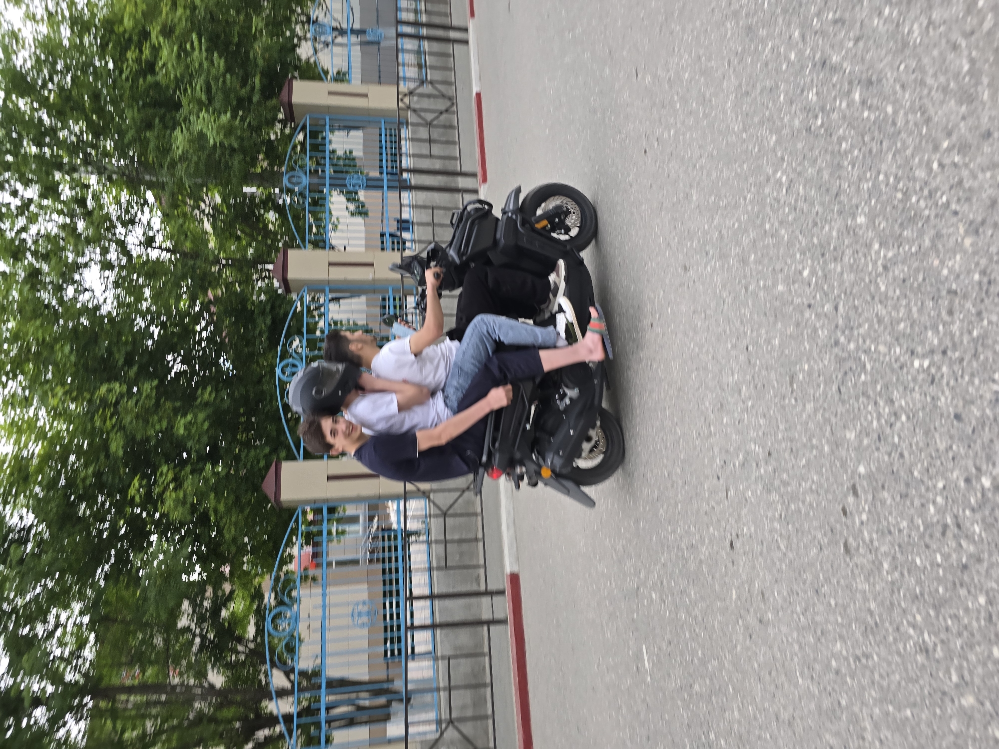 Scene: The image depicts two individuals riding a black scooter on a gray asphalt road. The scooter is positioned next to a blue metal fence with decorative circular designs. A red and white curb borders the road. Trees are visible in the background.
People: There are two individuals on the scooter. The driver is wearing a dark shirt and a helmet. The passenger is wearing a white shirt and blue jeans.
Objects: A black scooter, a blue metal fence, an asphalt road, trees, and a helmet are visible.
Actions: Two individuals are riding a scooter on a road.
Environment: The scene appears to be outdoors, possibly in a residential or urban area.