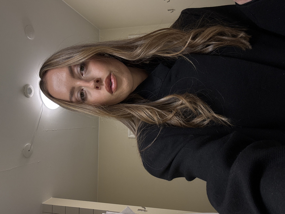 A young woman with light brown hair and fair skin poses indoors. She wears a black top. Her hair is styled with waves and highlights. The background consists of a plain wall with overhead lighting fixtures and tiles below. The image is taken at a slight angle, with the woman positioned towards the upper right.