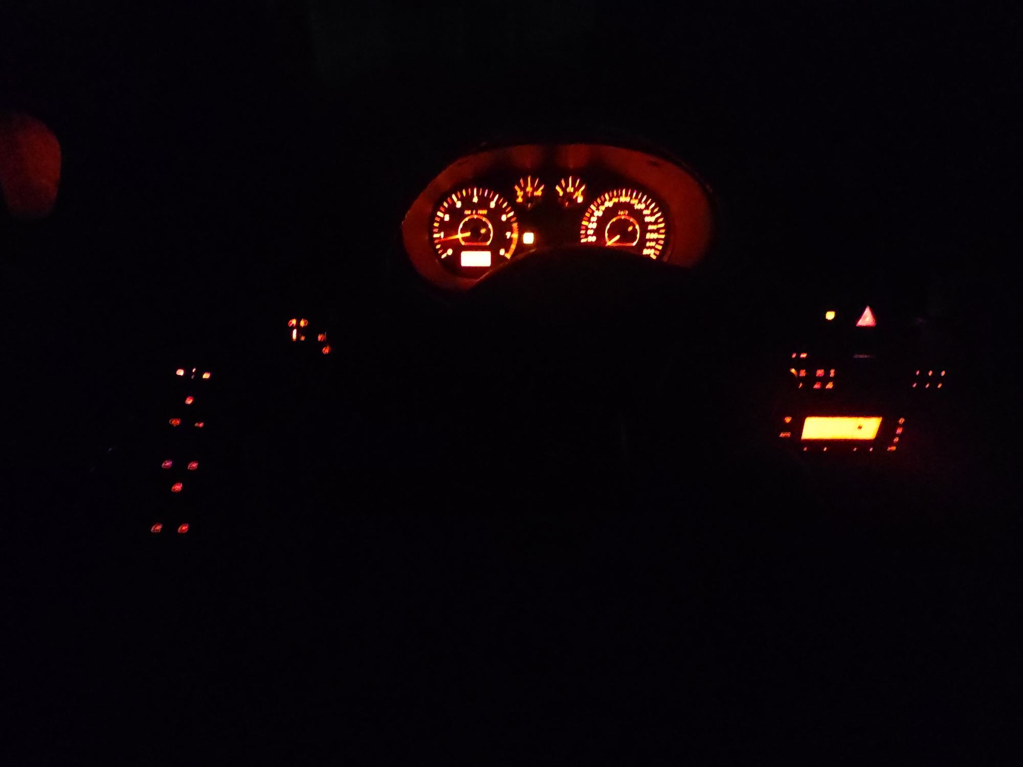 The image shows the illuminated dashboard of a car at night. The dashboard features a speedometer, RPM gauge, and other warning lights, all glowing in shades of red and orange. To the right of the dashboard is the car's radio, its screen also glowing orange. Various buttons with red indicator lights are visible on either side of the steering wheel. Due to the lack of detail and visual cues in the image, and the extreme darkness, it is impossible to determine the location as Sofia, Bulgaria. There are no people, specific objects or interactions visible that would allow for identification of geographic area.