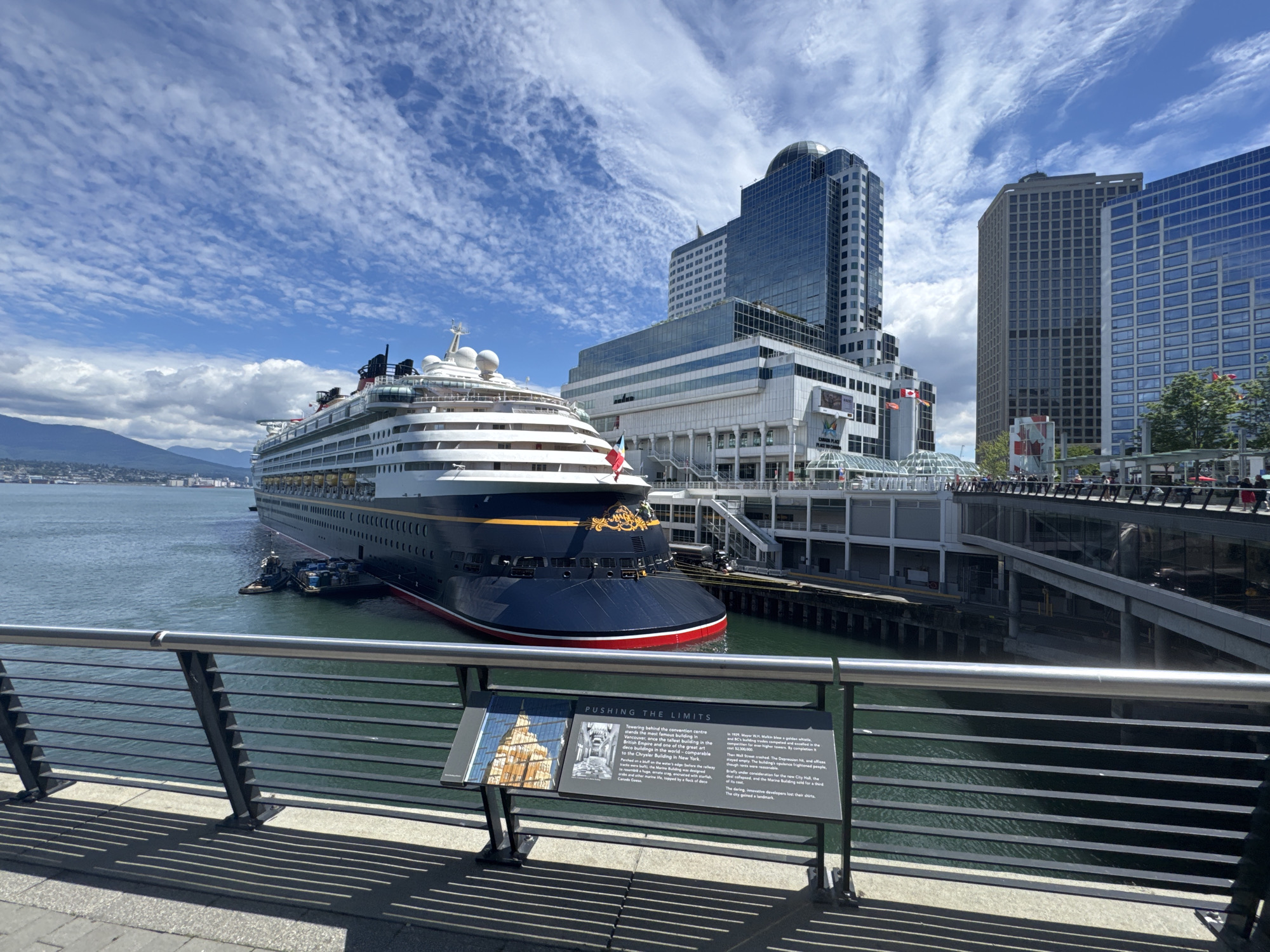 The image captures a view of Canada Place in Vancouver, Canada, on a bright, partly cloudy day. A large cruise ship, the Disney Wonder, is docked at the waterfront. The ship, predominantly navy blue with white decks and red trim, has its iconic yellow funnel with Mickey Mouse ears visible. Several smaller tugboats are positioned near the ship's bow.
In the background, the distinctive sail-like roof structures of Canada Place are visible, along with several modern glass-fronted skyscrapers that dominate the Vancouver skyline. These buildings represent the commercial and business district of the city. The water in the foreground is a vibrant turquoise color.
In the immediate foreground, there is a metal railing and a black metal sign with white text and images. The sign appears to be an informational plaque, with the title "PUSHING THE LIMITS" clearly legible. The plaque provides historical context about the surrounding buildings, including the convention center and older structures in the city. The ground in the foreground is paved with grey concrete slabs, with shadows cast by the railing onto the walkway. The overall scene conveys a bustling urban waterfront environment with a prominent cruise ship as the central subject.