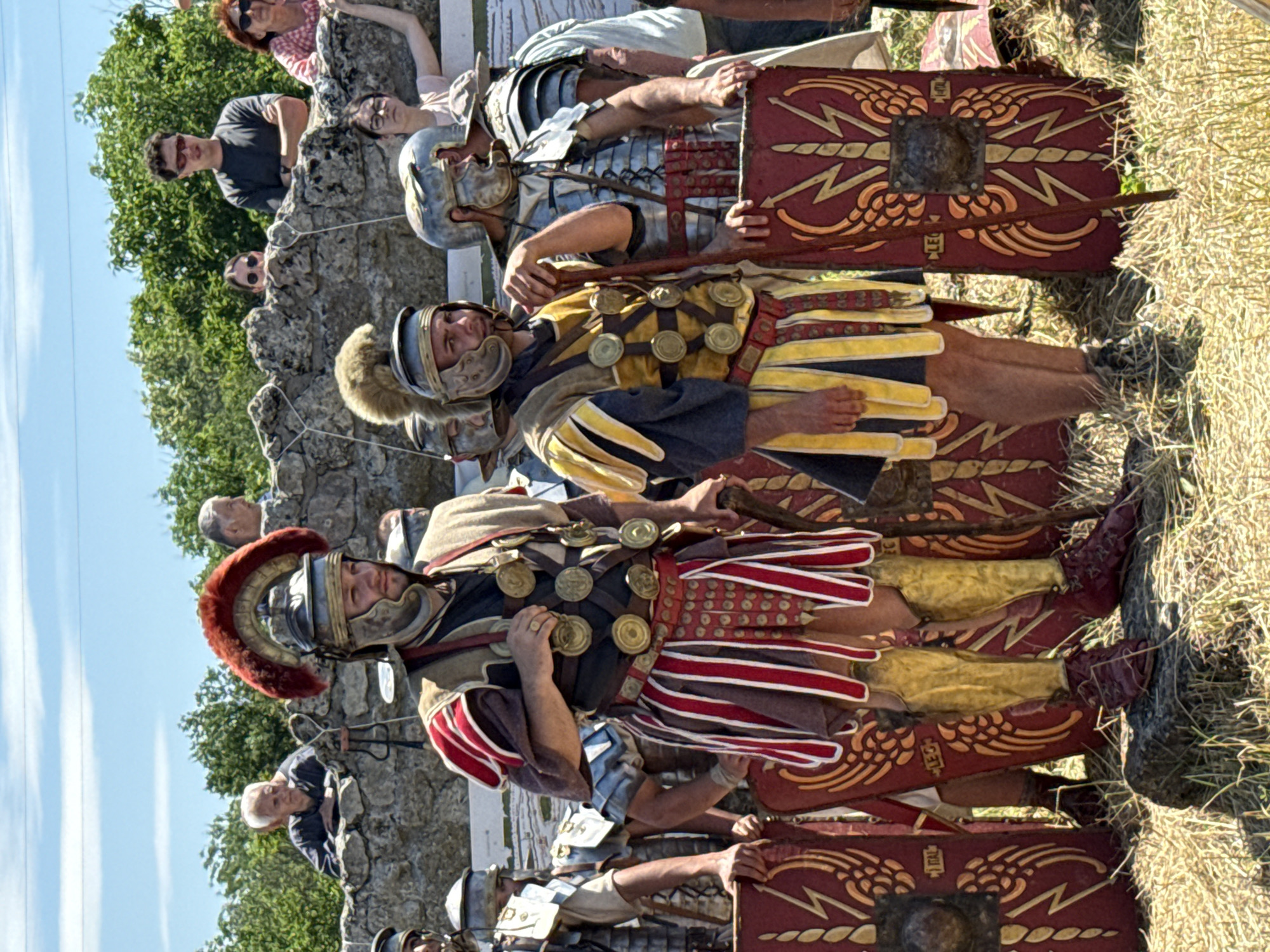 Gigen, Bulgaria. The scene captures a historical re-enactment featuring Roman legionaries outdoors under a clear blue sky. Eight individuals, dressed in various forms of Roman military attire, are positioned in the foreground and midground on dry, straw-colored grass. Two central figures are prominent. The figure to the left wears a helmet with a distinct red plumed crest, a dark tunic with red and brown striped pteruges, golden greaves, and red caligae, with multiple gold disc-shaped phalerae adorning his chest straps. He holds a wooden staff in his right hand. To his right, a second central figure wears a helmet with a brown fur-like crest, a dark tunic with yellow and blue striped pteruges, golden greaves, and red caligae, also displaying gold phalerae. He holds a wooden staff in his left hand. Both central figures have red rectangular scuta (shields) leaning beside them, adorned with golden winged lightning bolt motifs and central bronze bosses; one visible shield features the inscription 'ITAL' and another 'LEG'. Additional re-enactors in lorica segmentata armor and helmets are partially visible to the left and right of the central figures, also equipped with shields. In the background, behind a low, roughly constructed grey stone wall, five spectators in contemporary casual clothing observe the event. These include an older man with white hair, a man with dark hair and sunglasses, a woman with dark hair and sunglasses, another woman with red hair and sunglasses, and a young woman with glasses. Green trees and bushes form the natural backdrop behind the spectators and the stone wall. The ground in the immediate foreground consists of dry, light brown grass and some darker earth or rock formations. The scene is illuminated by bright natural sunlight.