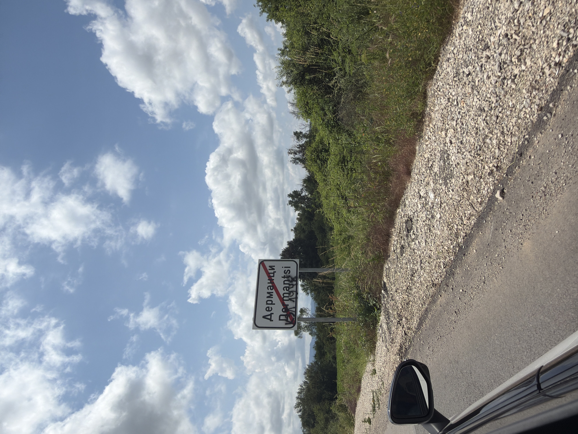 The image displays a daytime outdoor scene captured from the perspective of a vehicle, likely rotating 90 degrees clockwise from its intended landscape orientation. The sky is bright blue with scattered white cumulus clouds, suggesting fair weather.
The scene features a rural road winding through lush green vegetation. On the right side, a paved asphalt road transitions to a gravel shoulder, and then to overgrown grass and bushes. A densely forested or heavily vegetated area lines the horizon, with trees and shrubs prominent.
A white rectangular road sign, indicative of leaving a populated area, stands beside the road in the middle ground. The sign has a black border and features text in both Cyrillic and Latin scripts. The Cyrillic text reads "Дерманци," and the Latin text below it reads "Dermantsi." A thick red diagonal line crosses through both names, signaling that the vehicle is exiting the settlement of Dermantsi.
In the lower right corner, a black car side mirror and a portion of the car's window frame are visible, confirming the vantage point from inside a moving vehicle. The overall impression is of driving through the Bulgarian countryside, passing the sign indicating the end of the village of Dermantsi.