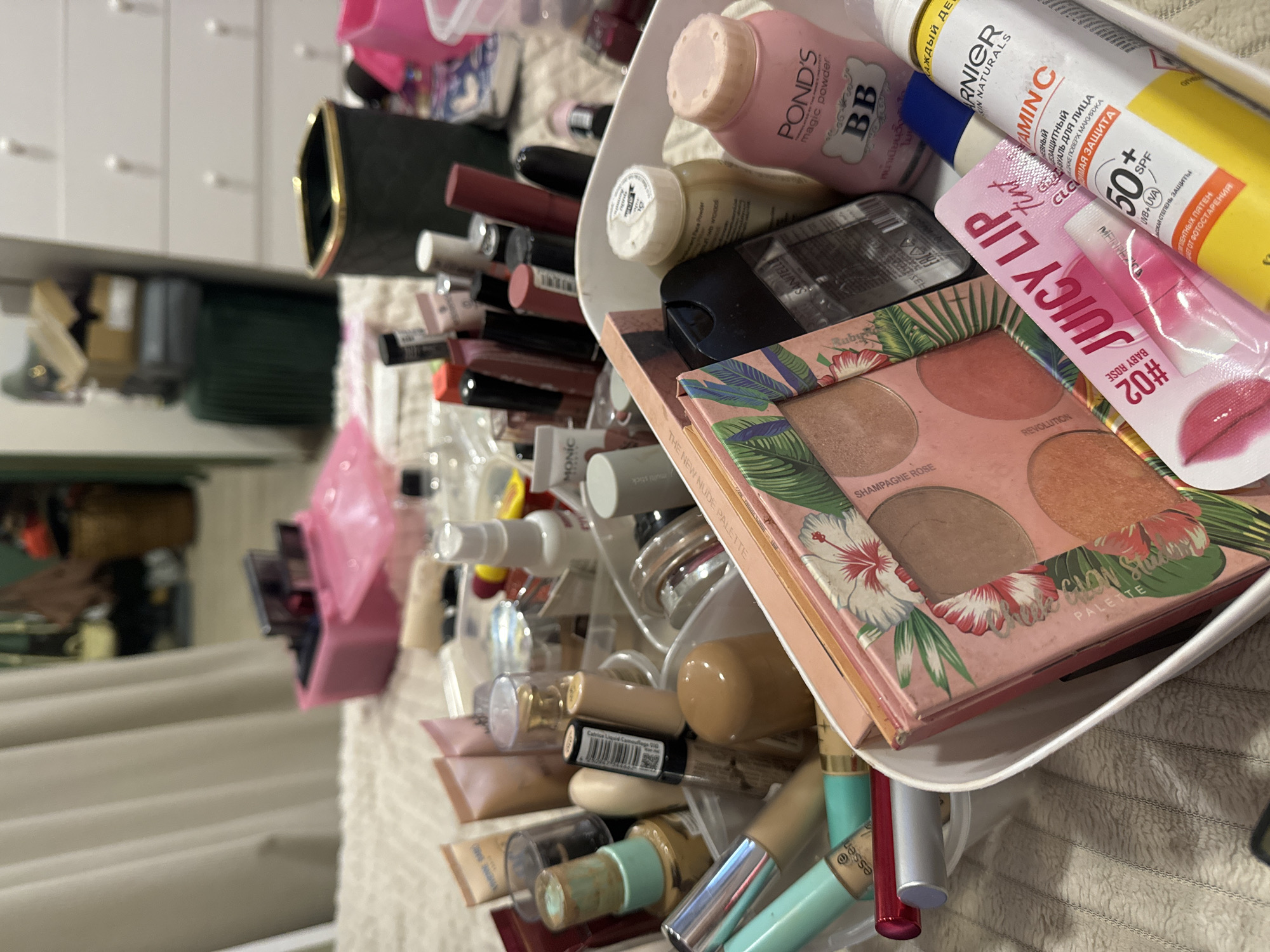 This image, rotated 90 degrees counter-clockwise, displays a cluttered collection of cosmetic and beauty products. The primary subjects are various makeup and skincare items, mostly contained within a white plastic organizer tray, with additional products scattered around it. The setting appears to be a personal space, such as a dressing table or vanity, with a white textured fabric (possibly a towel or blanket) beneath the items. In the blurred background, a white dresser or cabinet with multiple small handles and a mirror reflecting part of a room are visible.
No specific activity or event is taking place; it's a still life arrangement of personal care items. The lighting suggests an indoor setting, and no people are present.
Notable details include the variety of products, ranging from foundations and concealers to lip products, powders, and eyeshadows.
Visible text includes:
* On a pink bottle: "POND'S magic powder BB", "หน้าเนียนใส" (Thai for smooth, clear face).
* On a yellow and white tube: "GARNIER SKIN NATURALS", "VITAMIN C", "УВЛАЖНЯЮЩИЙ ЗАЩИТНЫЙ ФЛЮИД ДЛЯ ЛИЦА", "ДАЖЕ ПОВЕРХ МАКИЯЖА", "МАКСИМАЯ ЗАЩИТА", "50+ SPF UVB+UVA", "ОТ ПИГМЕНТНЫХ ПЯТЕН", "ЗАЩИЩАЕТ ОТ ФОТОСТАРЕНИЯ" (all Russian text relating to moisturizing protective fluid, high protection, and anti-aging).
* On a pink pouch: "JUICY LIP tint", "#02 BABY ROSE", "MENPELO".
* On a rectangular palette with tropical design: "REVOLUTION", "CHAMPAGNE ROSE", "PALETTE", "GLOW PALETTE" (partially visible).
* On a bottle: "Catrice Liquid Camouflage HQ".
* On a light blue tube: "essen" (likely essence).
* On a white stick product: "MONIC multi stick".
* On a small bottle: "SANITELLE".
* Partially visible on a box: "THE NEW NUDE PALETTE".