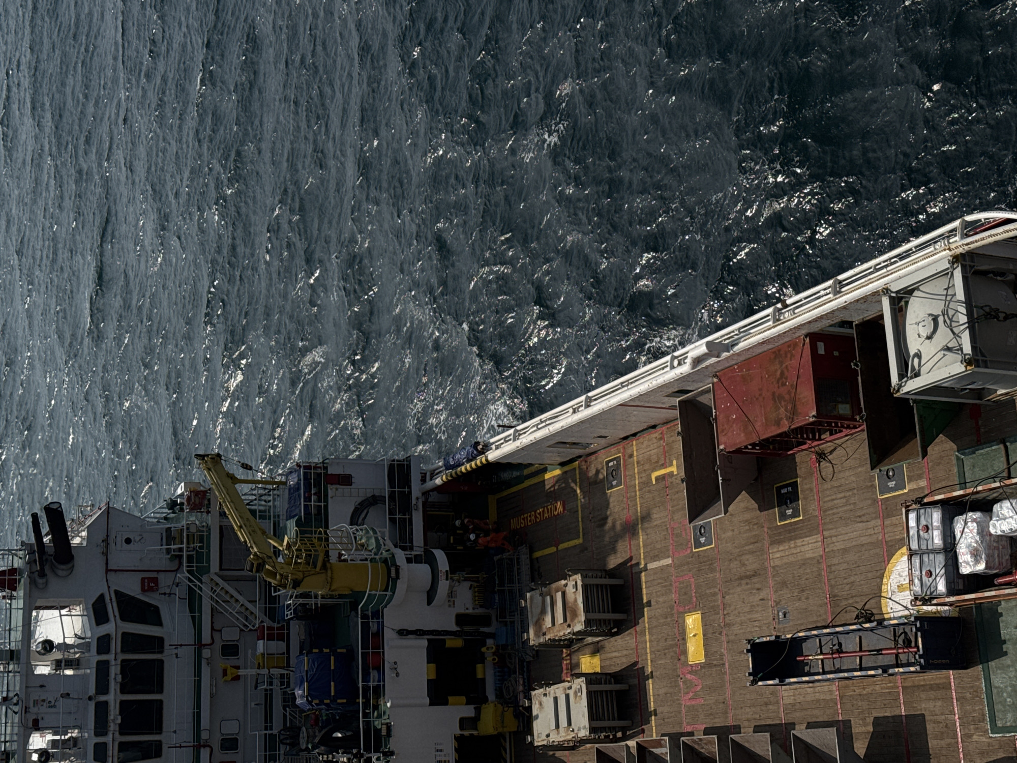 An elevated, medium-range, eye-level shot shows the deck of a white and gray vessel on dark, choppy ocean water. The ship's structure dominates the left side of the frame, featuring multiple windows, a yellow crane, and a white railing along the deck. On the deck, several metallic containers of various sizes are visible, along with red and yellow markings on the wooden surface. Two individuals wearing orange jumpsuits are observed near the ship's superstructure. The ocean, a deep blue-gray, is filled with visible waves and sunlight glinting off the water's surface.