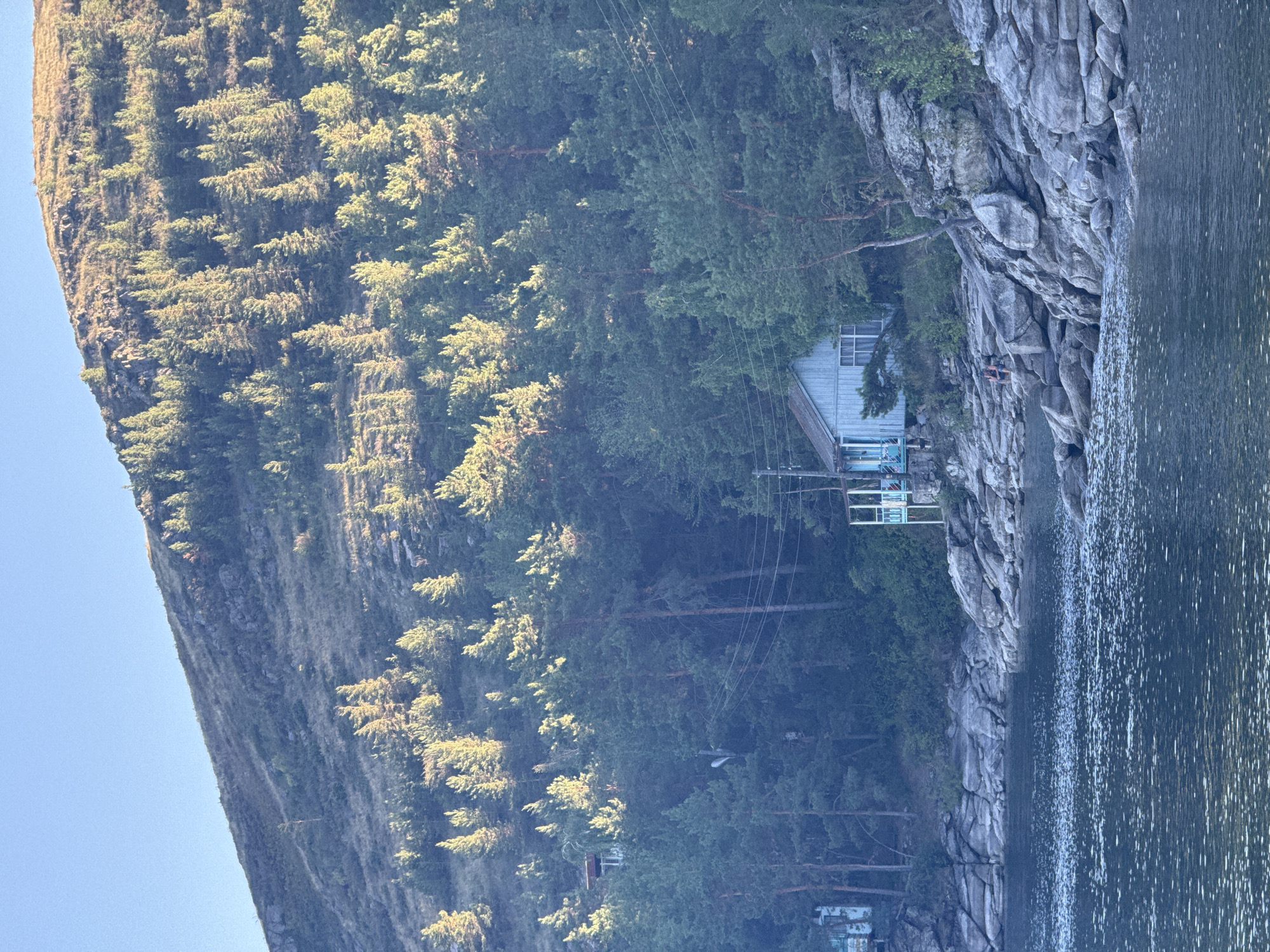 A body of water, likely a lake or river, fills the foreground of the image, its surface reflecting the light with ripples. Along the shoreline, a rocky embankment is visible, with large grey stones and patches of green vegetation. Nestled on this embankment, slightly elevated and partially obscured by trees, is a small, light blue wooden house with white trim and a porch. The house appears to be a rustic dwelling, possibly a cabin or a summer home.
Behind the house, a dense forest of pine trees covers the slopes of a hill. The trees are a deep green, with some areas catching the sunlight and appearing golden. The hill rises steeply, its upper reaches rocky and sparsely vegetated, transitioning into a lighter, sun-drenched slope at the very top. The sky above is a clear, pale blue. The overall scene depicts a serene natural landscape with a solitary dwelling integrated into the environment. There are two individuals sitting on the rocks near the house.