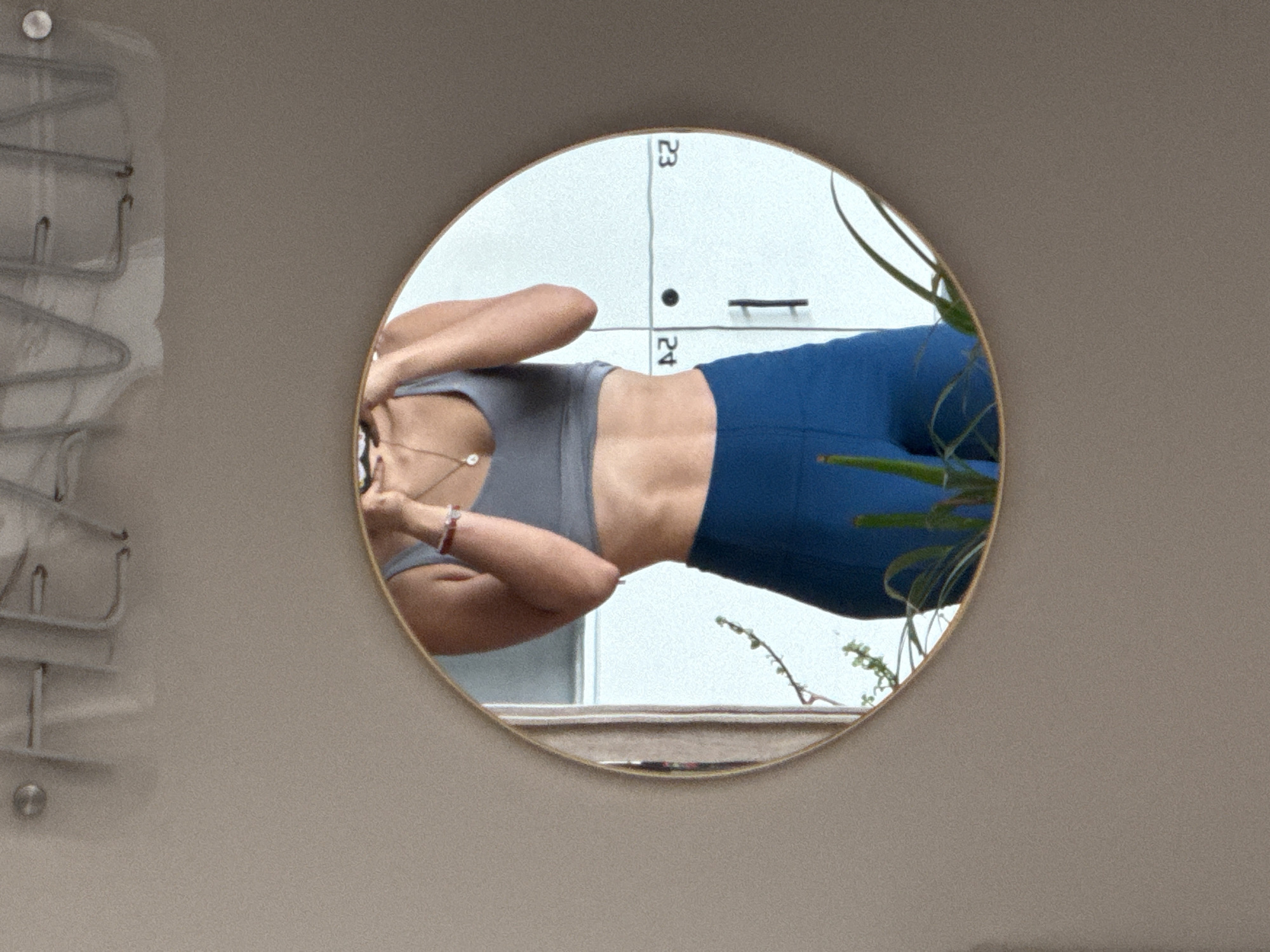 The image displays an indoor mirror selfie, oriented such that the subject appears lying on their side. A person, presumably female, is reflected in a round mirror with a thin, light-colored frame, located in Puebla, Mexico. The person is wearing a grey sports bra and blue leggings, holding a phone with both hands to take the picture. Their midsection is toned, and a delicate necklace with a pearl-like pendant is visible around their neck, along with a red or pink bracelet on one wrist.
The reflection behind the person reveals a white surface resembling lockers or storage cabinets, marked with black dividing lines and the numbers "23" and "24." A dark handle or hook is also visible on this white surface. Green foliage from a plant partially obscures the right side of the reflection within the mirror. At the very bottom of the mirror's reflection, a light-colored horizontal surface, possibly a floor or ledge, is visible.
The mirror itself is mounted on a plain, light beige or taupe wall. To the left of the mirror, a light-colored metal rack with multiple curved hooks, likely for hanging items, is attached to the wall. A round, silver-toned fixture, possibly a screw head or decorative knob, is also visible on the wall near the rack. The overall setting, particularly the numbered storage units, suggests a gym, yoga studio, or changing room environment.