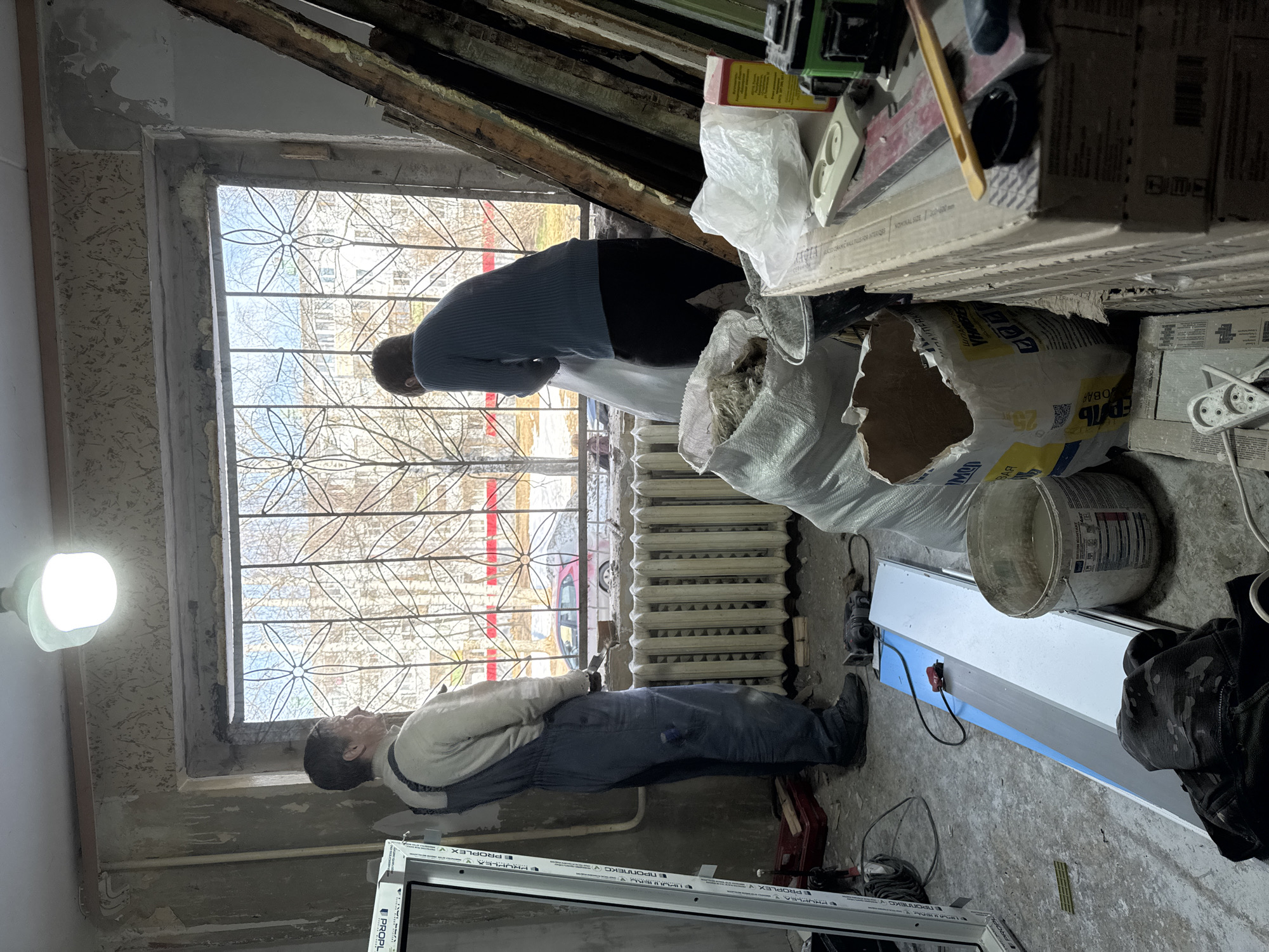 A wide-angle, indoor shot depicts what appears to be a construction or renovation project underway. Two individuals are present. One individual, dressed in a blue long-sleeved shirt and dark pants, appears to be working near the top of an old window frame. The second individual is seen wearing overalls. They are looking toward the window. The window has a metal decorative grid in front of the glass and is framed by older, possibly damaged, window frames. Outside, a view of leafless trees, buildings, and a red structure can be seen. In the foreground are several bags of materials, a bucket, tools, and what appears to be a new window frame wrapped in protective material. The overall setting suggests an interior space under renovation, likely in an older building. Based on metadata, this location is Chaikovsky, Russia.