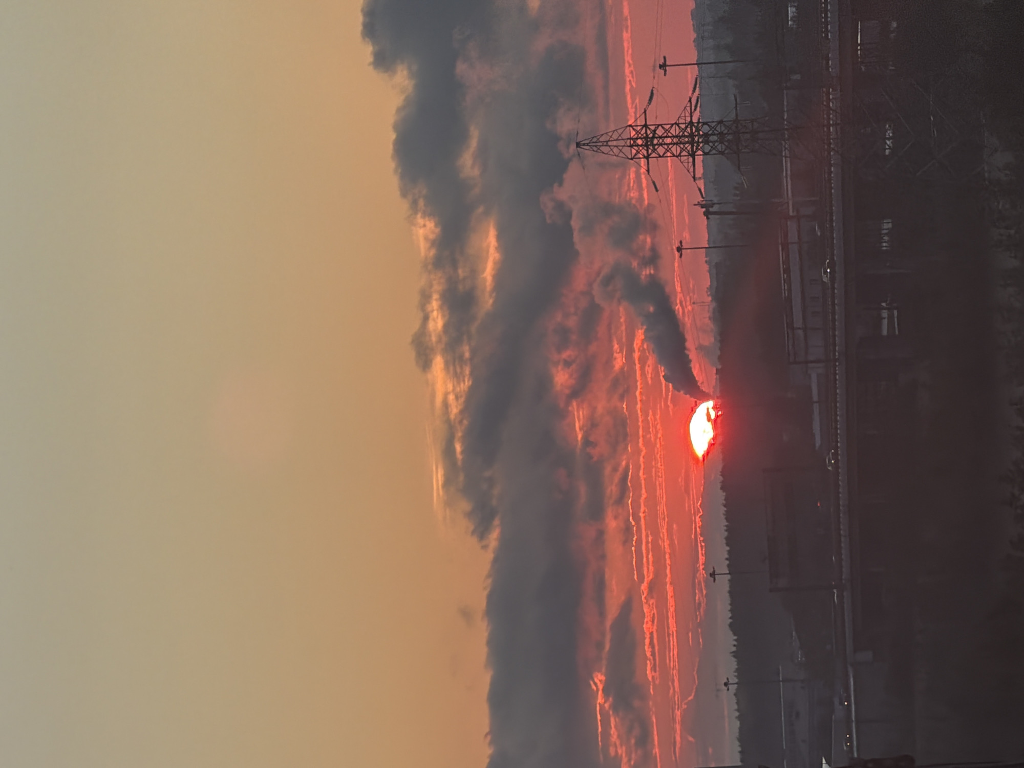 A vertical, medium shot captures an industrial landscape at sunset. The sky is a vibrant gradient of orange and pink, with dramatic, dark clouds streaked with red light. The sun, a bright red orb, is partially obscured by these clouds. In the mid-ground, a large, lattice-style electricity pylon stands prominently on the right. Below this, a multi-level highway structure is visible, with cars driving on it. To the left of the pylon, industrial buildings and trees form a dark silhouette against the colorful sky, with a plume of white smoke rising from a factory stack. The overall impression is one of urban development juxtaposed with natural phenomena.