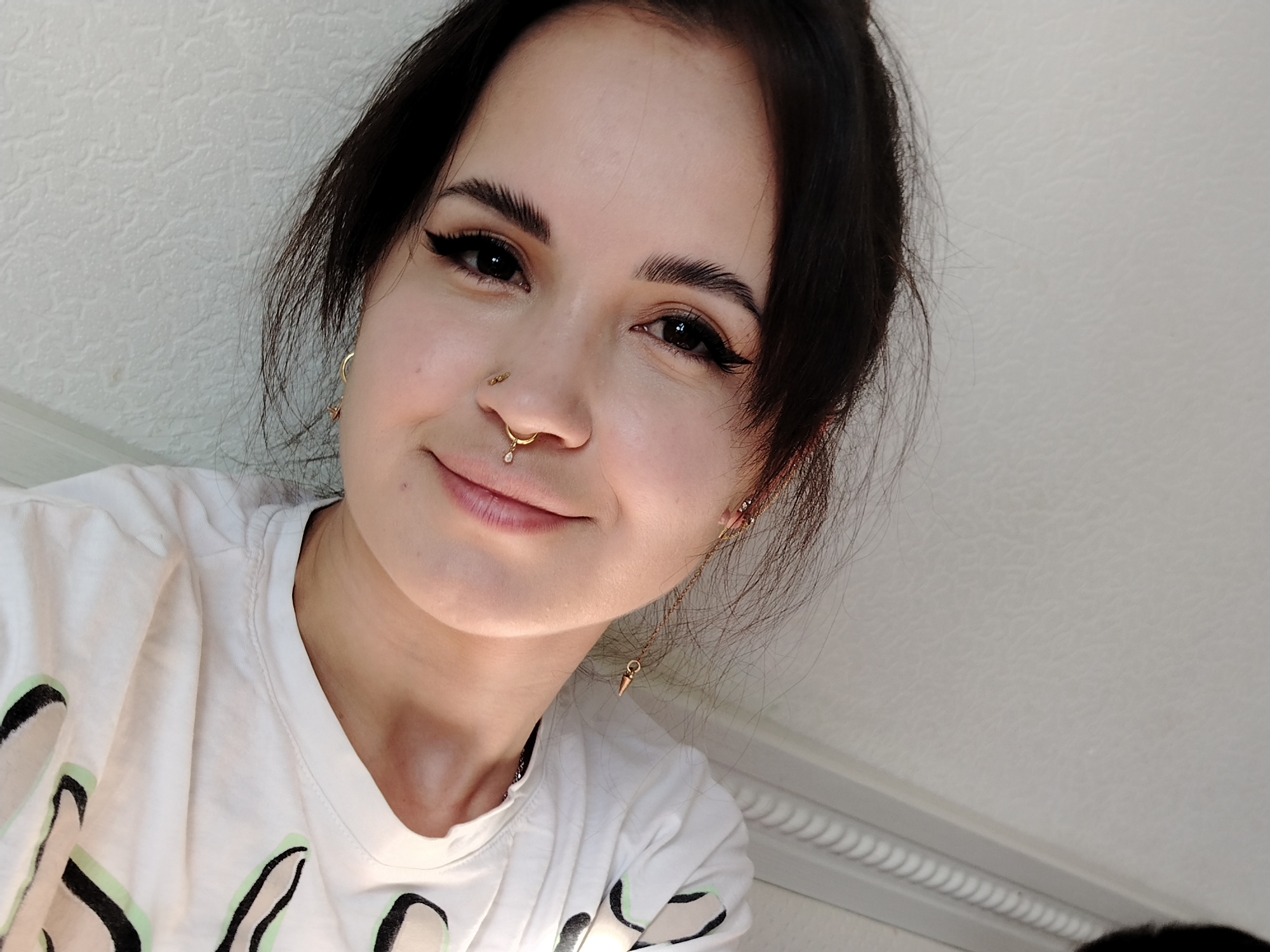A close-up selfie of a young woman with fair skin, dark brown hair, and visible makeup including winged eyeliner. She has a gold ring in her left nostril and a gold hoop earring with a dangling jewel in her right nostril. Additional gold earrings are visible in both ears. She is wearing a white T-shirt with black and green abstract designs. The background is a plain white wall. No evidence indicates the location is in Noviy, Russia.