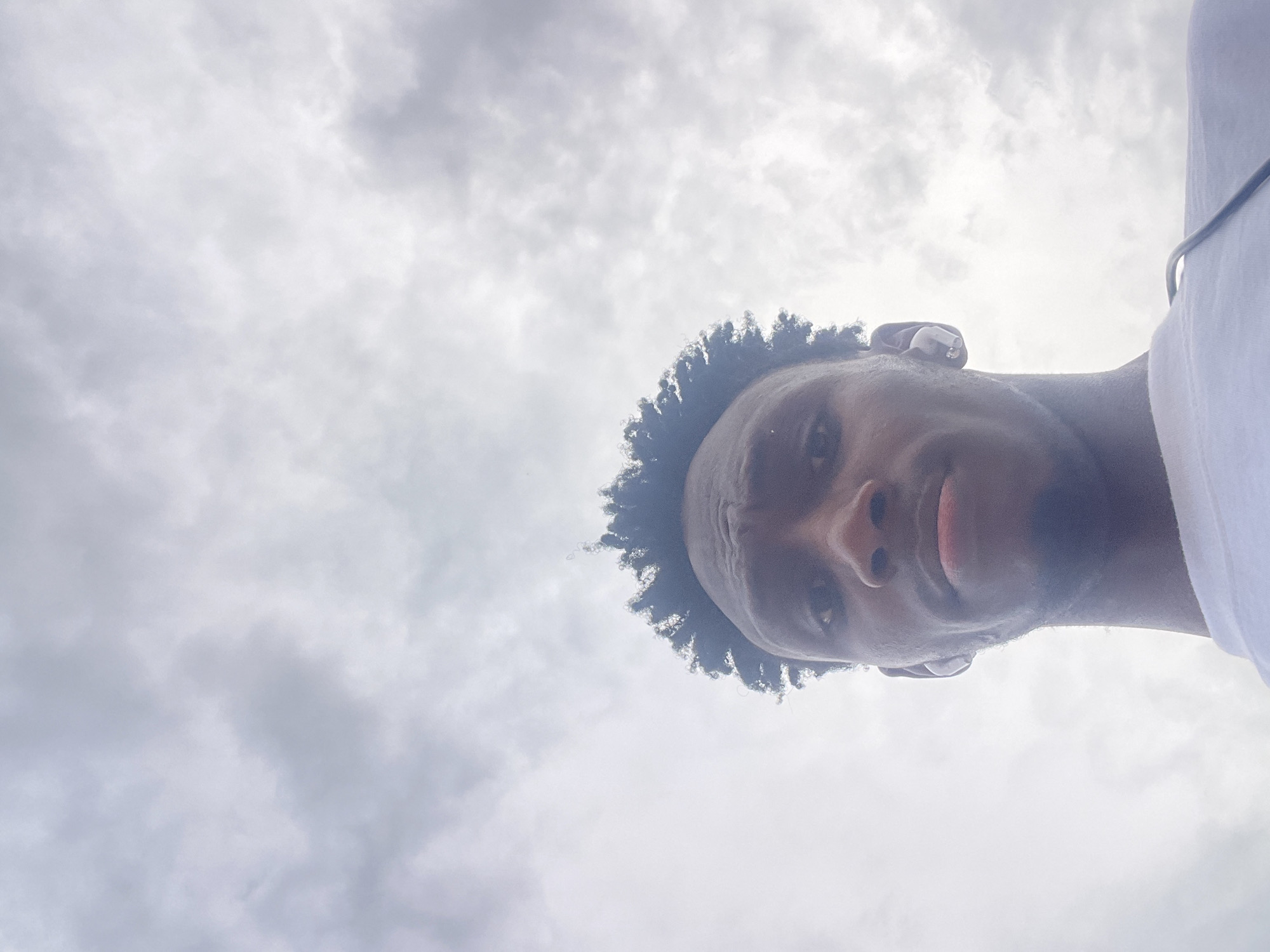 A low-angle portrait captures a young man looking directly downwards into the camera, set against an overcast sky. His head is significantly tilted to the left. He has dark skin, short, curly black hair, and is wearing a white top, the shoulder of which is visible on the right side of the frame. A white wireless earbud is visible in his left ear. His facial expression is serious and intense, with a slight frown and subtly parted lips. The background consists entirely of diffused gray and white clouds, suggesting an overcast daytime sky. There is no visible text. The location is contextually identified as Lokoja, Nigeria.