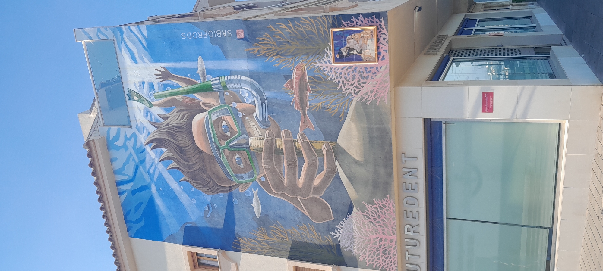 The image captures a vibrant street art mural on the facade of a commercial building in Manilva, Spain, under a bright, clear blue sky. The mural dominates the upper portion of the building, depicting an underwater scene.
The central subject of the mural is a large, stylized figure, possibly a man, with flowing brown hair, wearing a green snorkel mask and goggles. Bubbles surround his face, suggesting he is submerged in water. Intriguingly, he is actively brushing his teeth with an oversized yellow and white toothbrush. The underwater environment includes various fish, such as a reddish-brown fish and a smaller white one, along with intricate golden-brown seaweed and delicate pink and white coral structures. Rays of light penetrate the water from above, creating a dynamic and immersive effect.
Above the mural's main figure, the Instagram handle "@SABIOPRODS" is visible, identifying the artist or group responsible for the artwork. A small, framed tile art piece is embedded in the mural's upper right section, containing an abstract design and the faintly visible word "mousse."
Below the mural, the building's facade is modern, featuring large windows. The name "FUTUREDENT" is displayed vertically along the corner of the building, indicating it is likely a dental clinic. One of the lower windows features a red sign with the text "Allianz Asistencia" and a phone number "900 100 249." The bright daylight suggests it is daytime. The image is taken with a rotated orientation, making the vertical building appear horizontal.